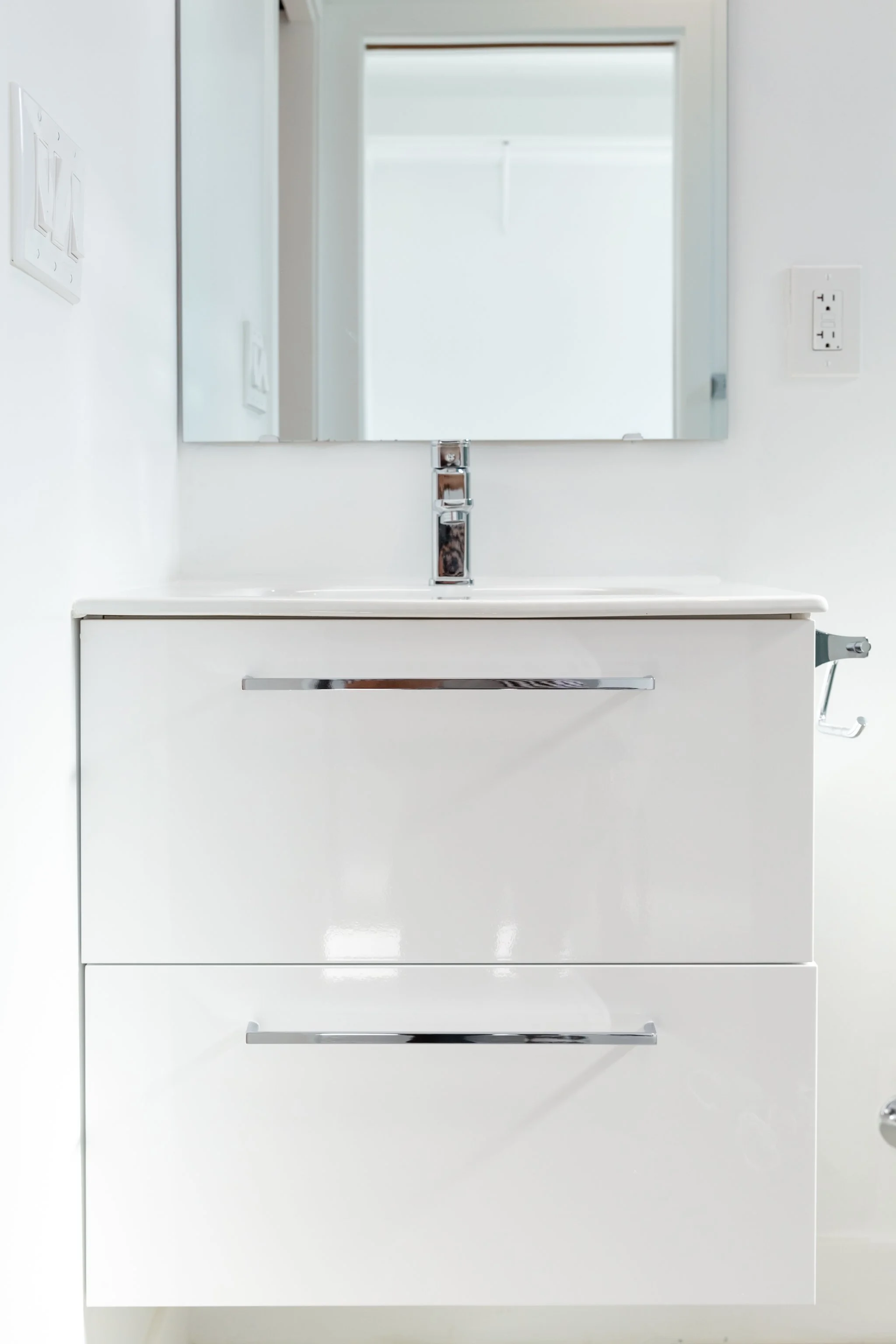 White bathroom vanity with a mirror, chrome handles, and a modern faucet, with electrical outlets on a white wall.