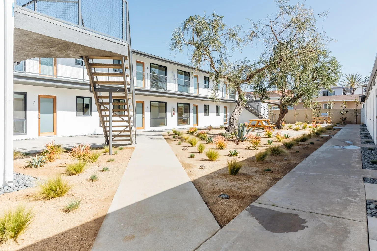 Apartment courtyard with two-story buildings, woven wood doors, and outdoor staircases, featuring desert landscaping with trees, succulents, and grass plants.