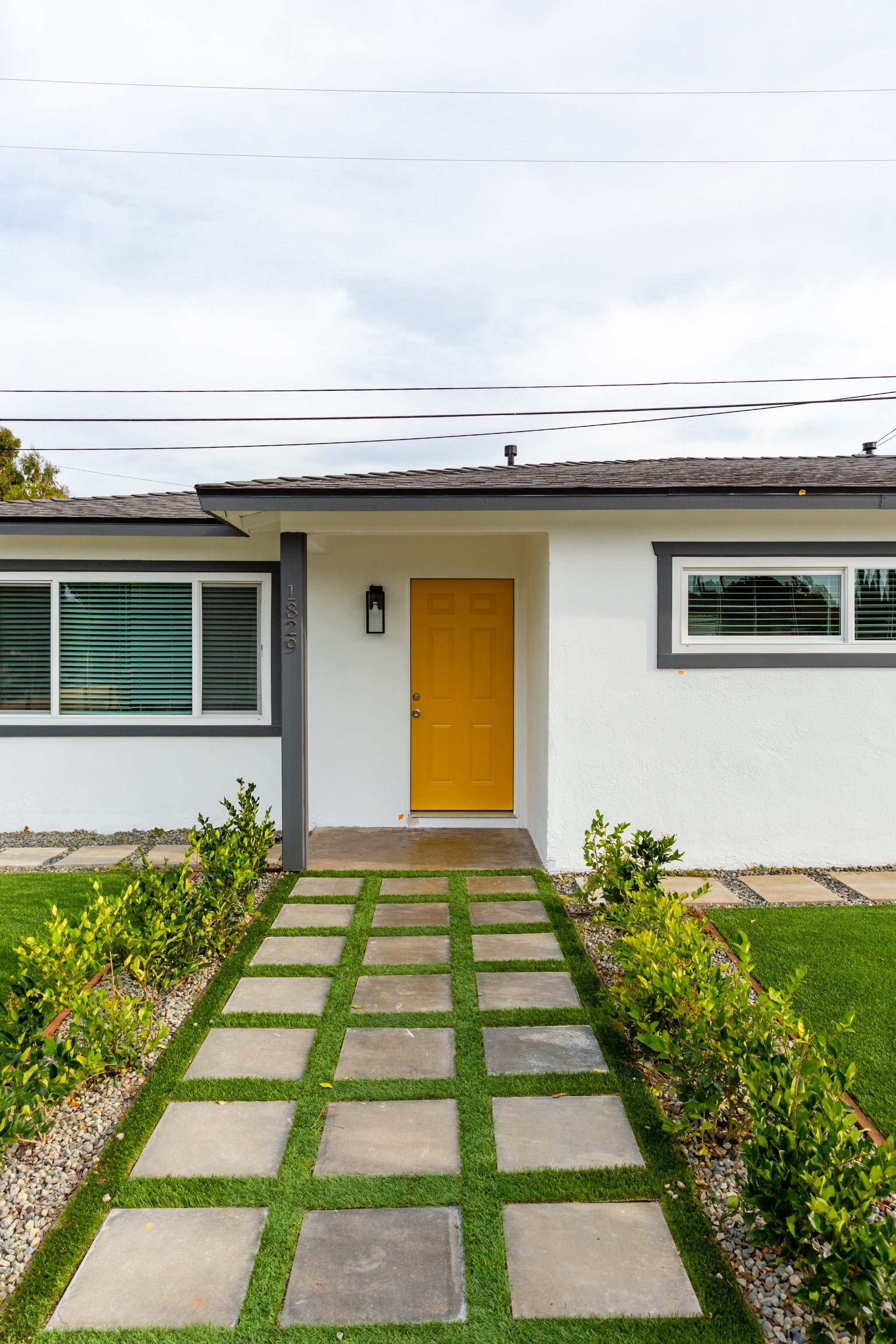 Front view of a modern house with a yellow door, large windows, and a small front lawn with a pathway made of concrete pavers and grass strips, surrounded by small bushes and landscaping rocks.