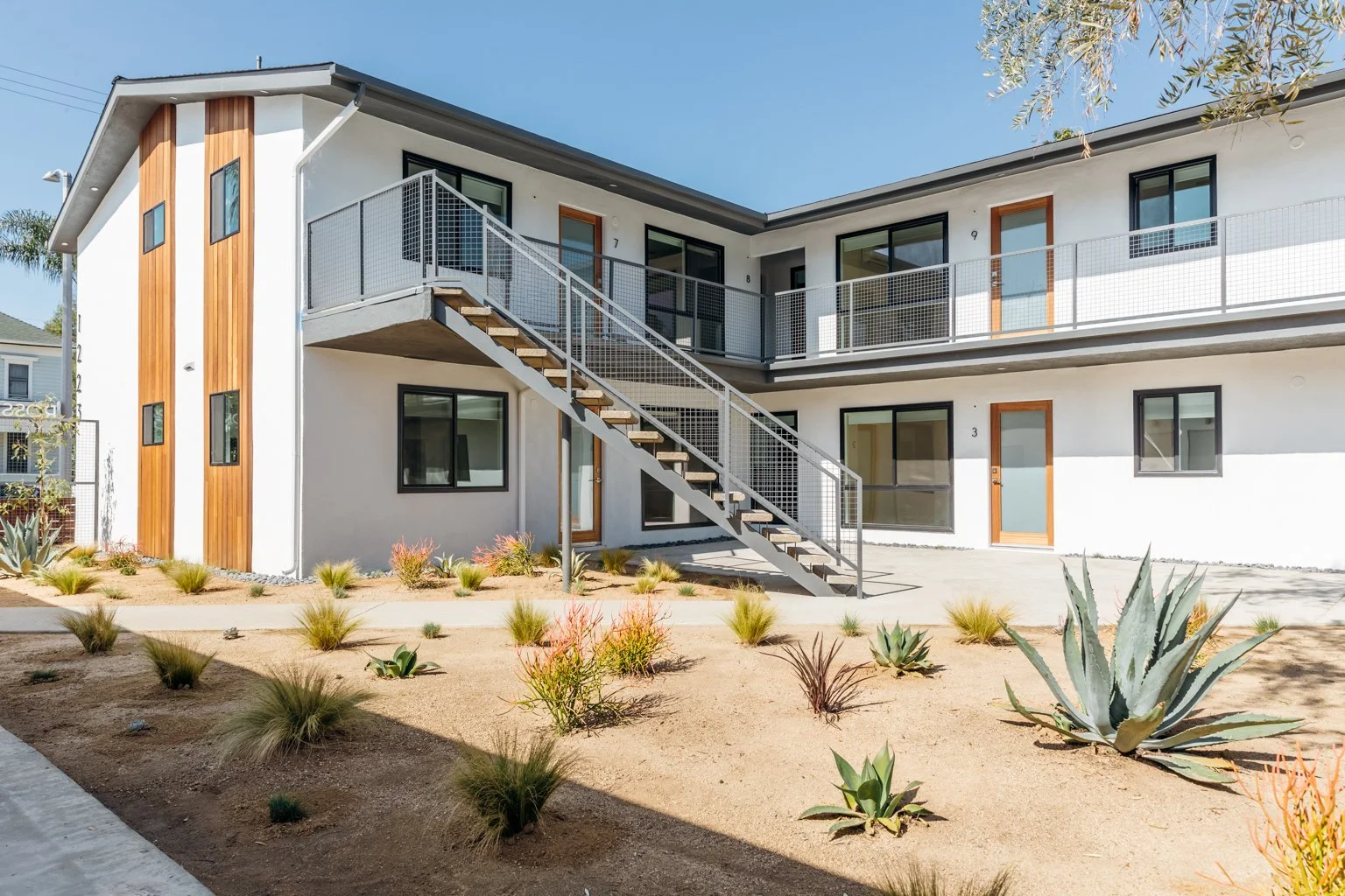 Modern two-story apartment building with white exterior walls, wooden accents, black window frames, and a metal staircase leading to the second floor. The front yard features desert landscaping with succulents and dry soil.