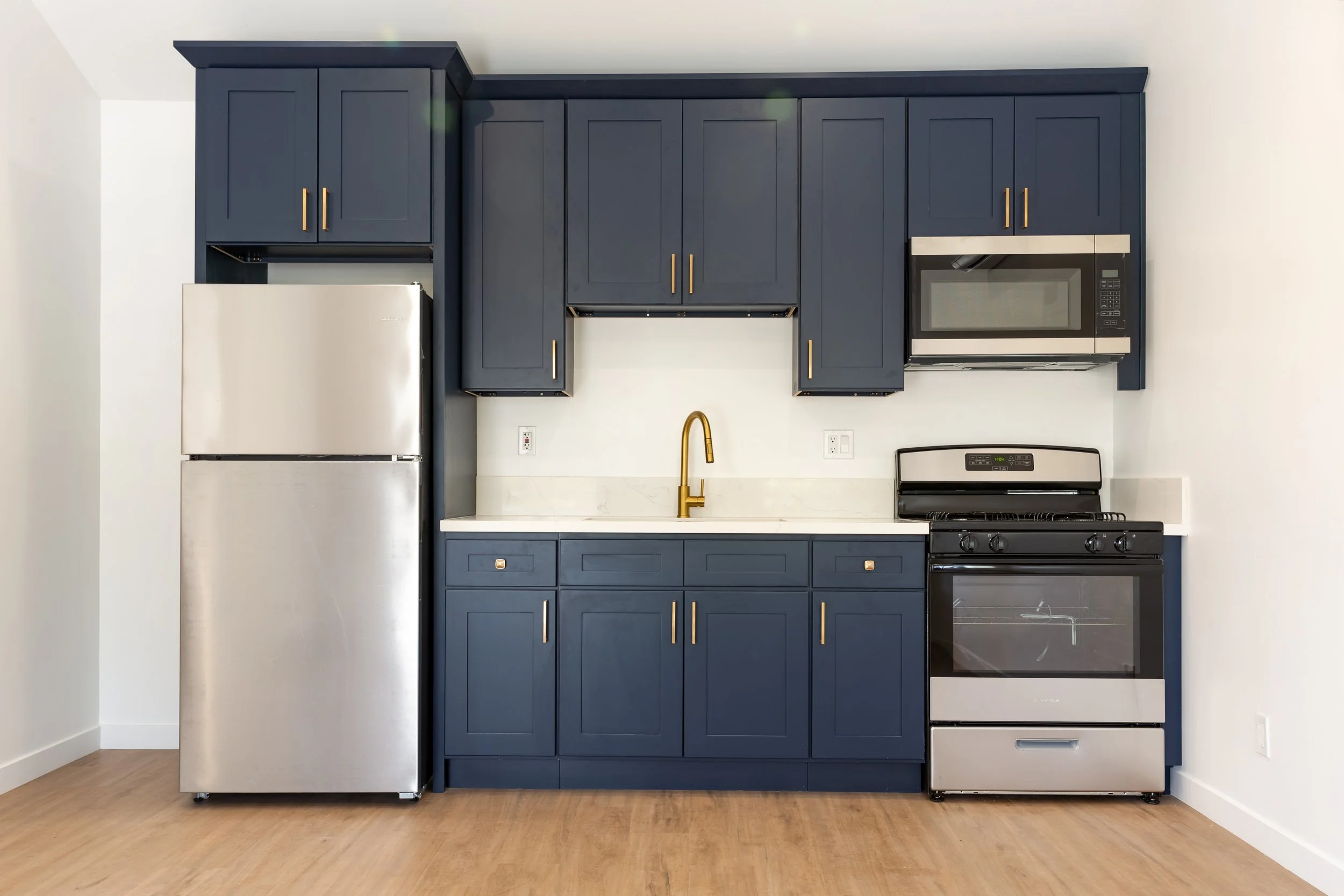 Modern kitchen with navy blue cabinets, stainless steel refrigerator, black stove with oven, microwave, white countertop, gold faucet, and hardwood flooring.