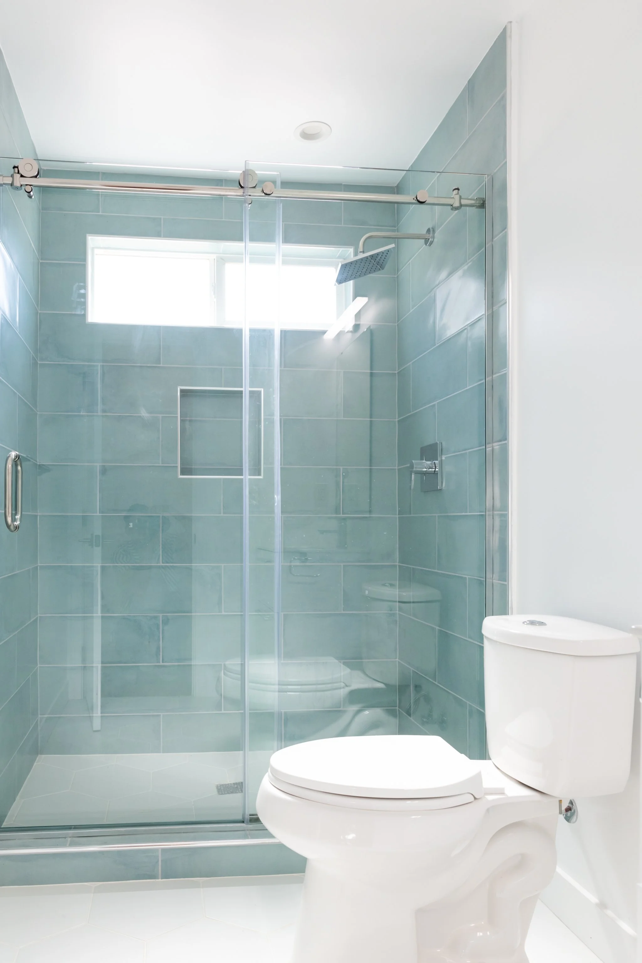 A modern bathroom with a glass-enclosed shower, a rain showerhead, light blue tiled walls, a white toilet, and a small window.