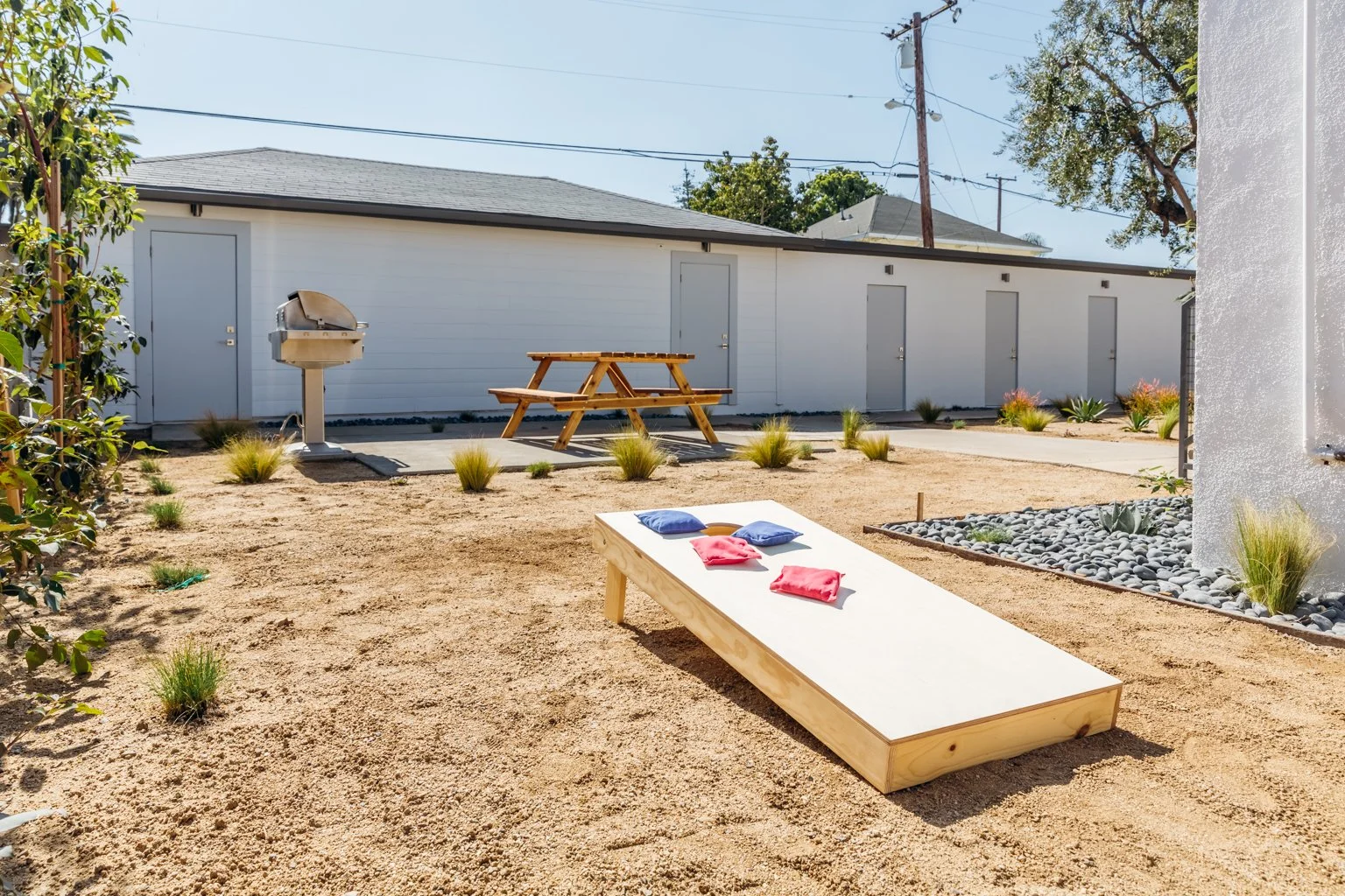 A backyard with a raised cornhole game, a picnic table, a grill, and a landscape with small plants and rocks, enclosed by a white shed and a white wall.