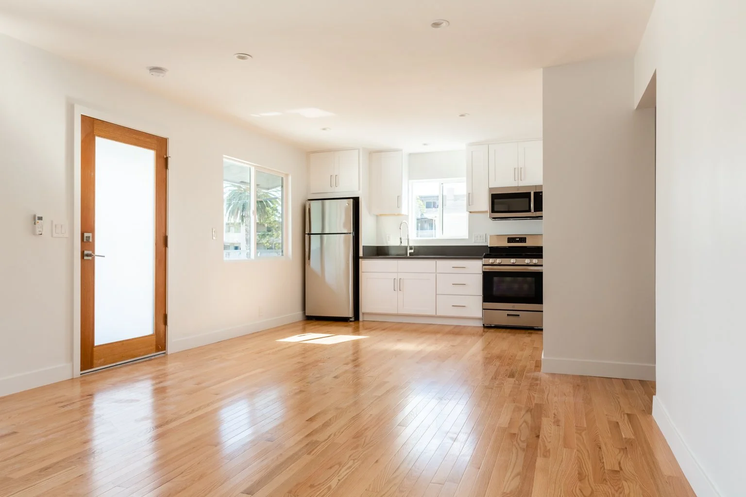 Empty kitchen and dining area with hardwood floors, white cabinets, stainless steel refrigerator, stove, microwave, and a window.