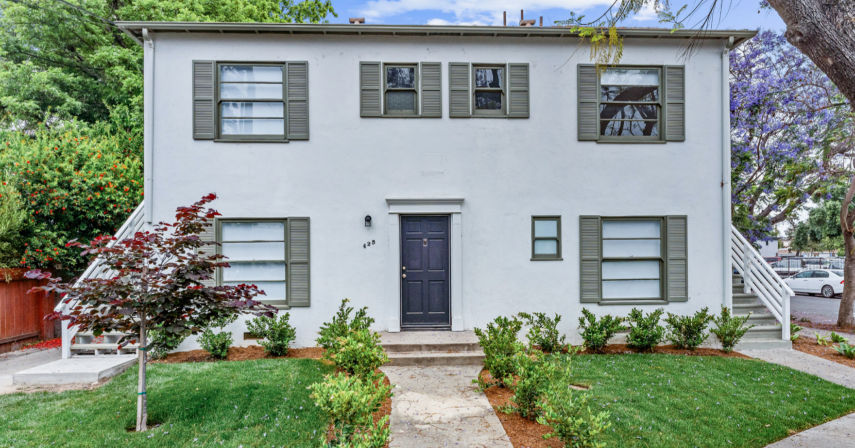 Two-story white residential building with external stairs on both sides, gray shutters on windows, and a black front door. Front yard with green grass, young trees, and small bushes, and parking lot with cars in background.