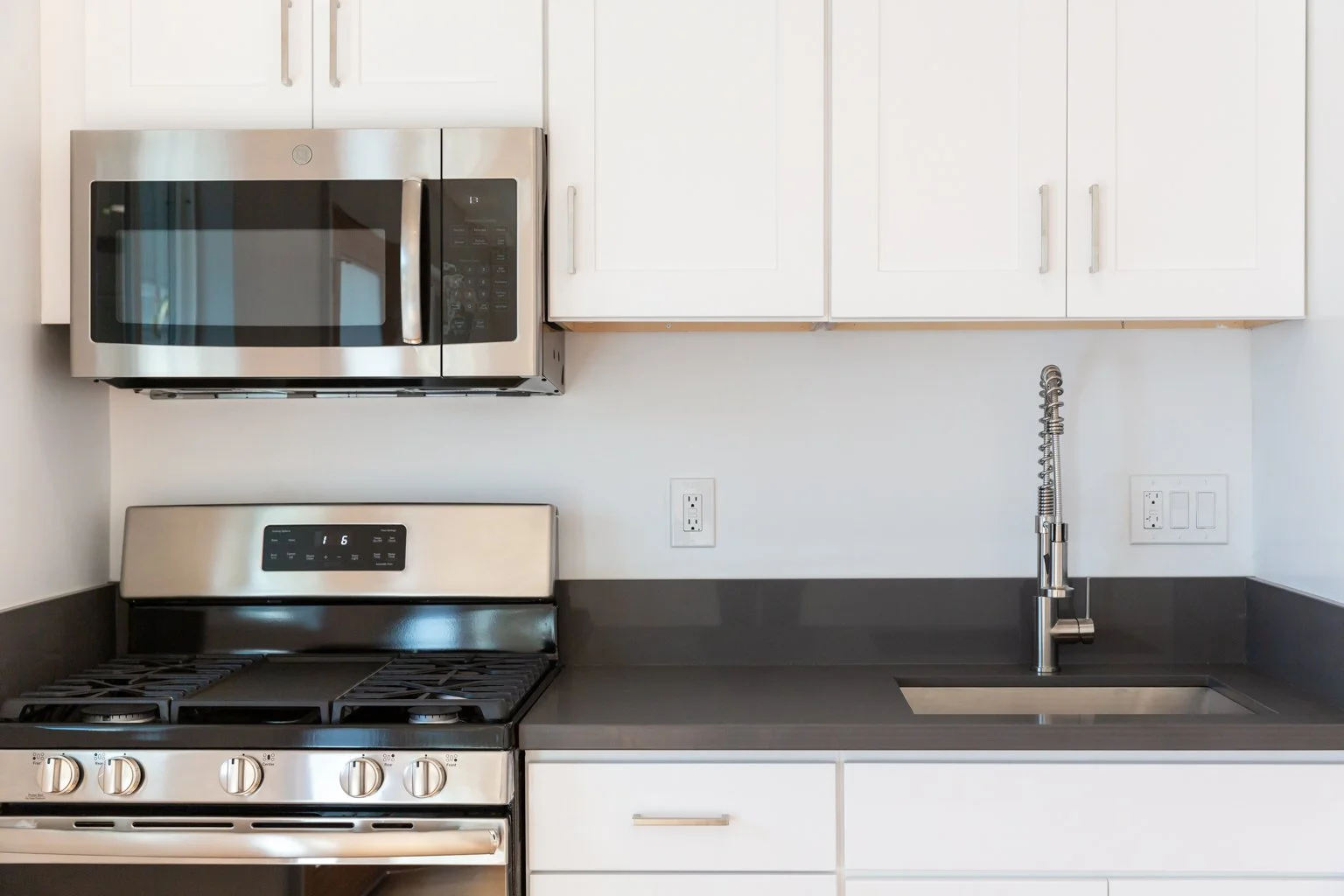 Modern kitchen with stainless steel microwave, stove, black countertop, white cabinets, and a silver sink with a spring-style faucet.