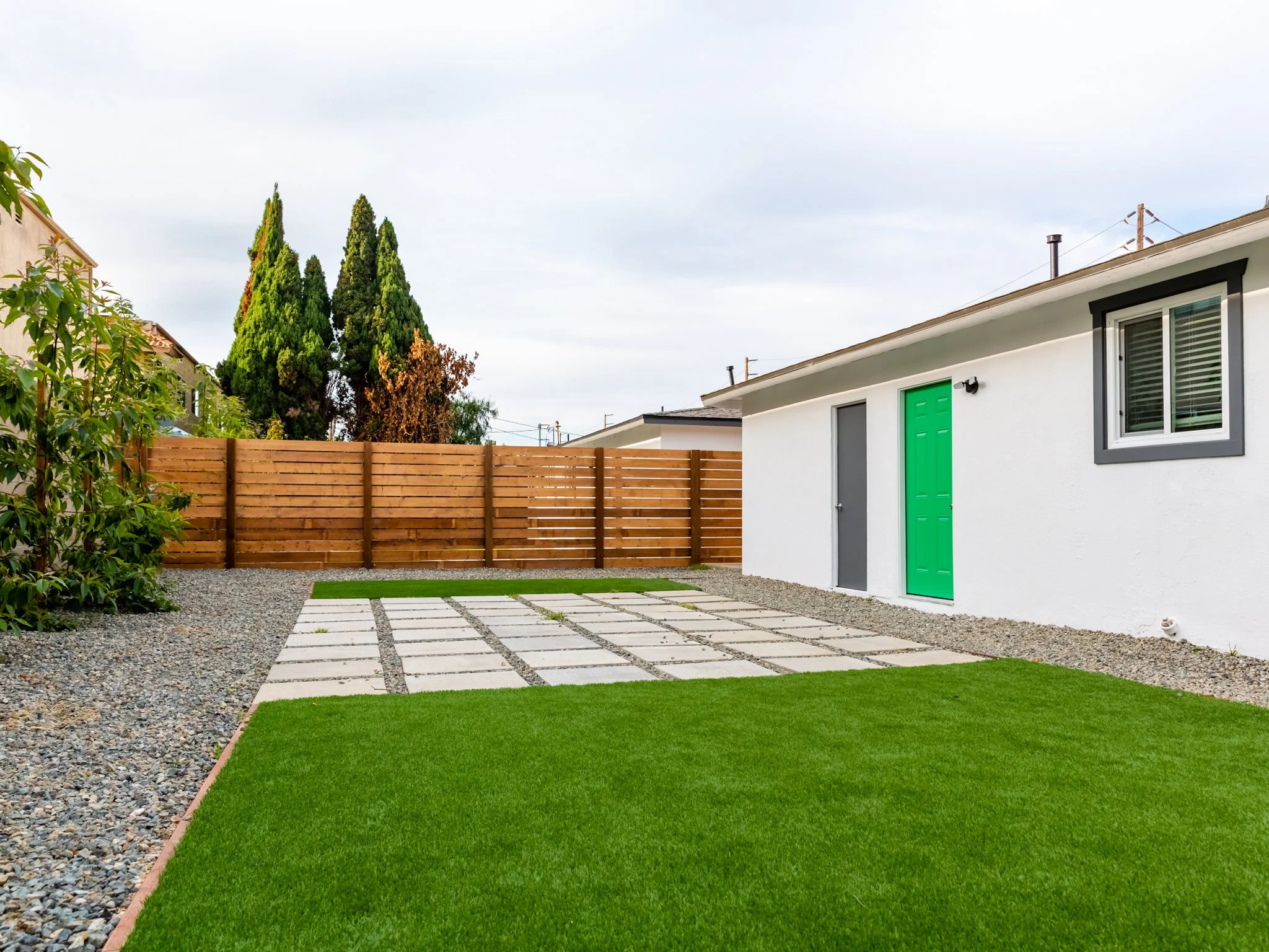 A backyard with a green lawn, gravel pathway, wooden fence, and a white house with a green door and a window. There are tall trees in the background under an overcast sky.