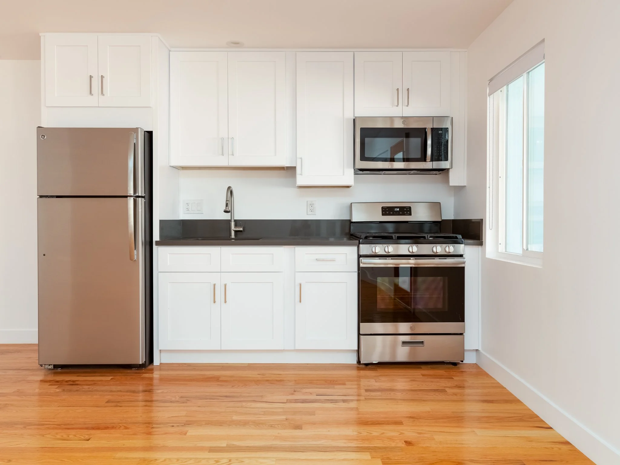 Modern kitchen with white cabinets, stainless steel refrigerator, microwave, and oven, along with a wooden floor and a window on the right.