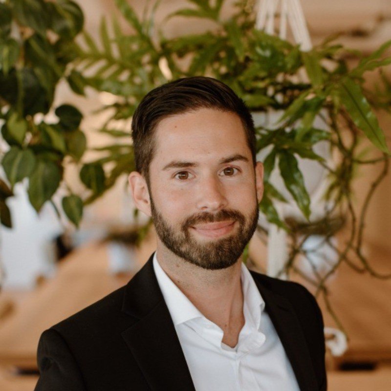 A man with dark hair and a beard, dressed in a black suit jacket and white shirt, standing indoors with green plants in the background.