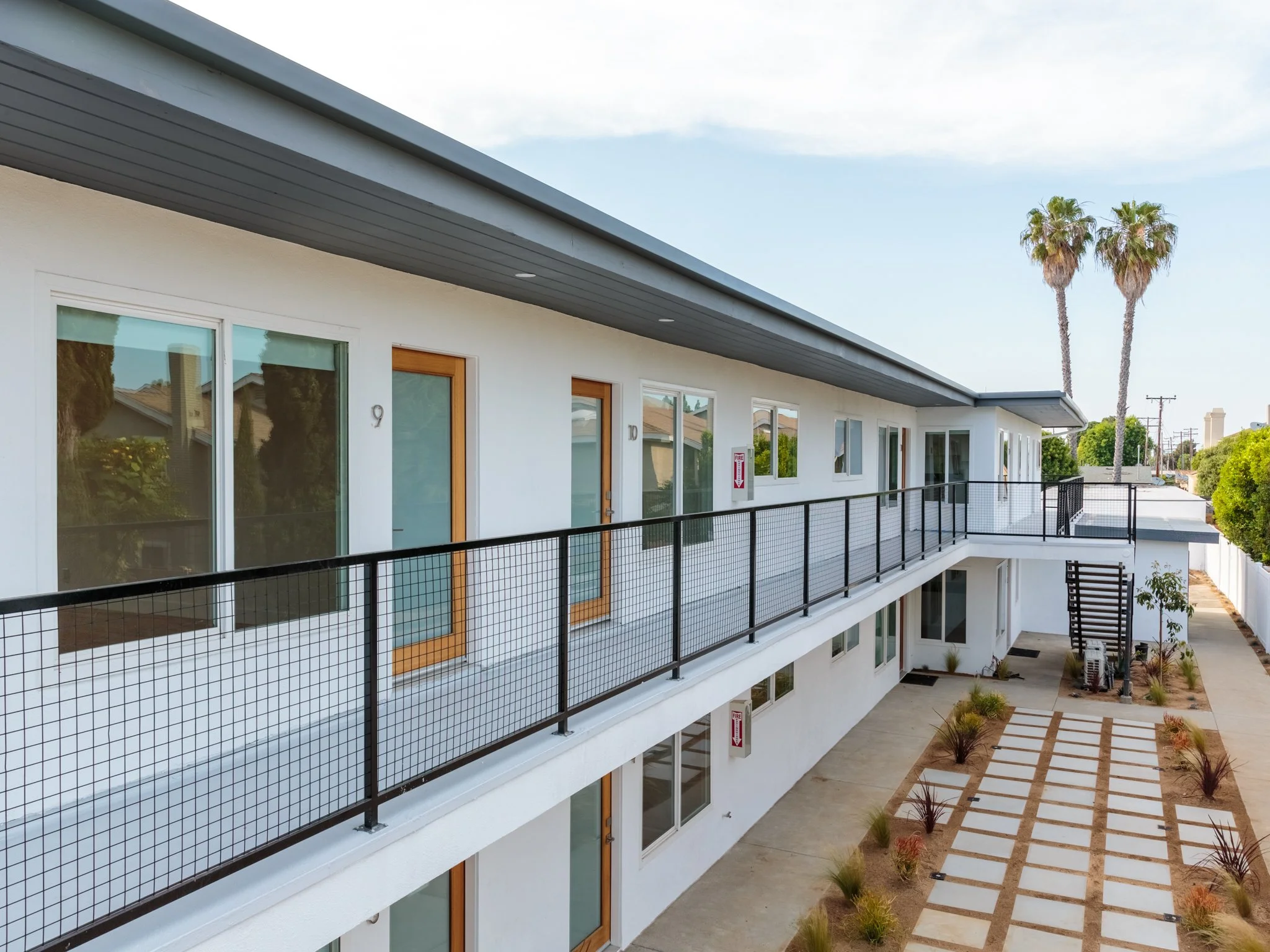 Modern white apartment building with large glass sliding doors, black metal railings, and a public outdoor area with patterned paving and small shrubbery plants. Two tall palm trees are visible in the background.