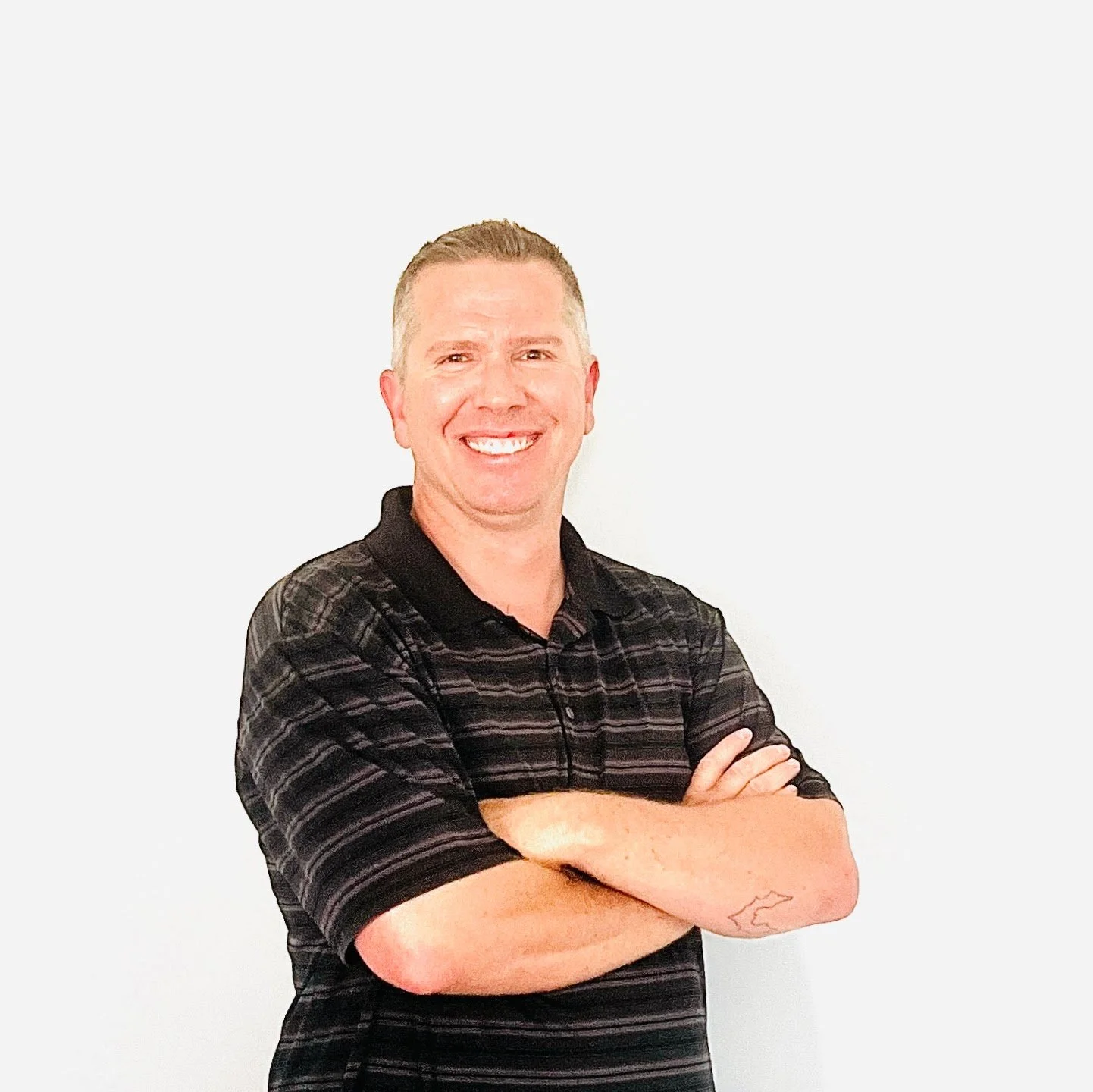 A smiling man with short light hair wearing a black and gray striped polo shirt, crossing his arms, standing against a plain white background.