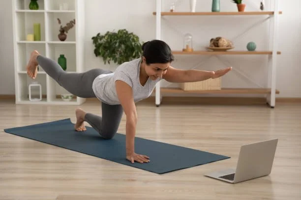Woman exercising on a yoga mat while following a workout video on a laptop in a home setting.