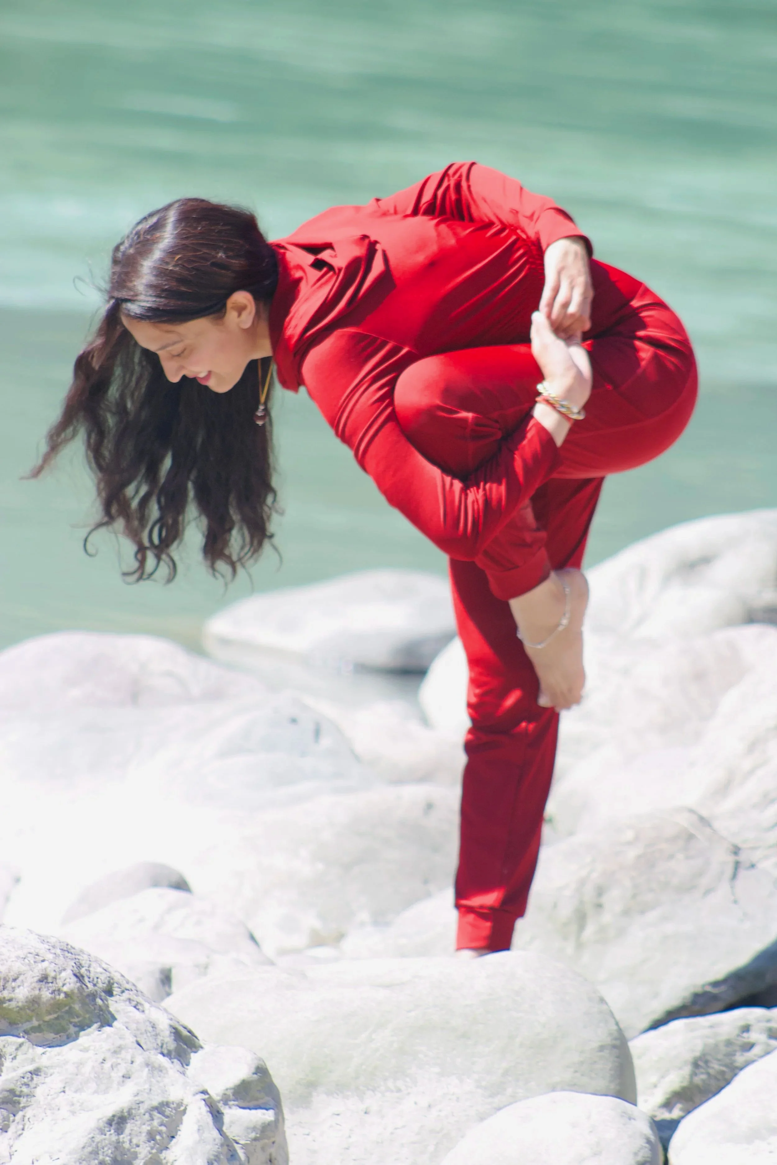 A woman dressed in a red tracksuit standing on rocks by a body of water, bending forward and smiling.