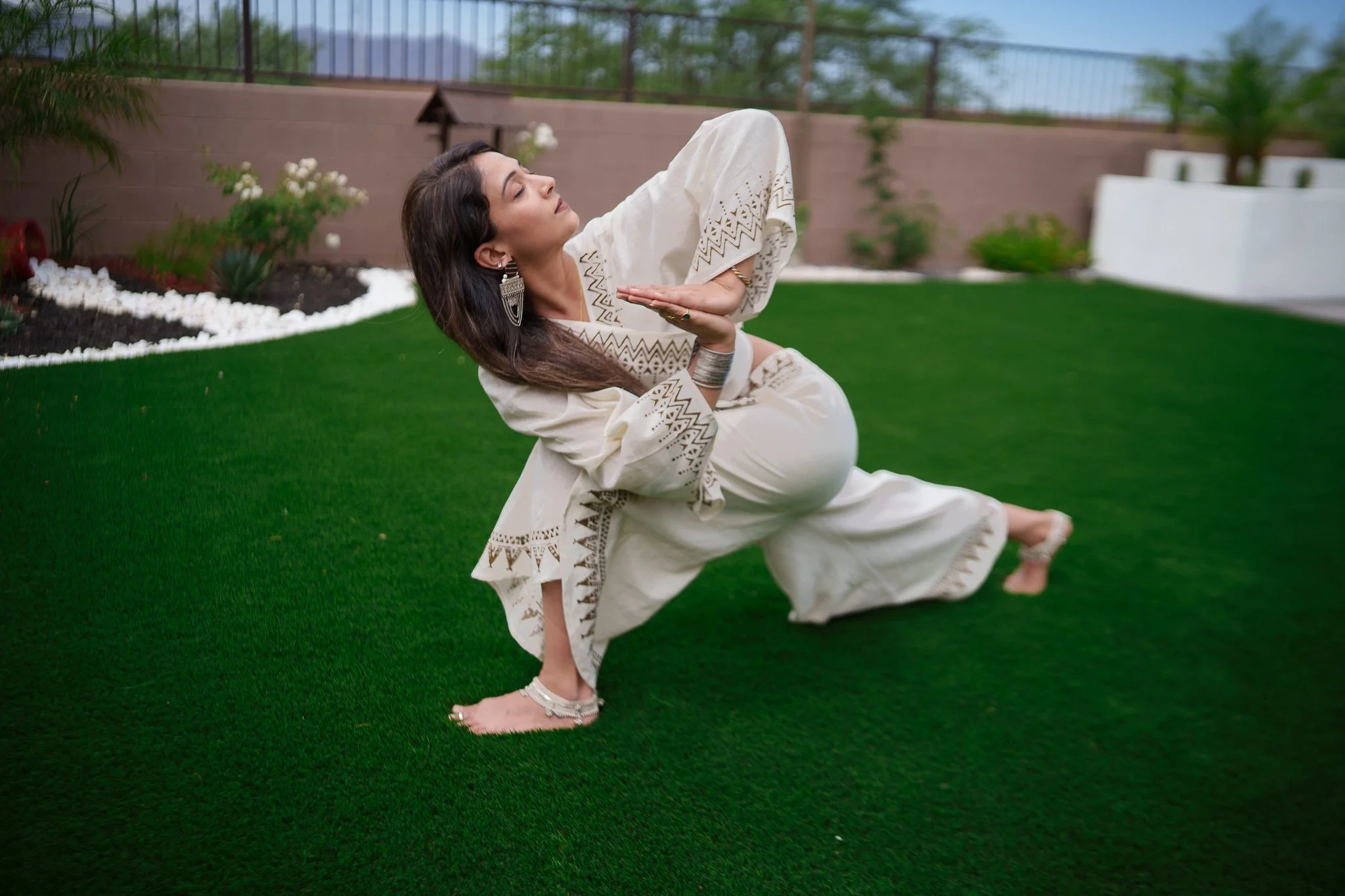 A woman in traditional Indian attire practicing yoga outdoors on a green lawn with flower beds and a brown wall in the background.