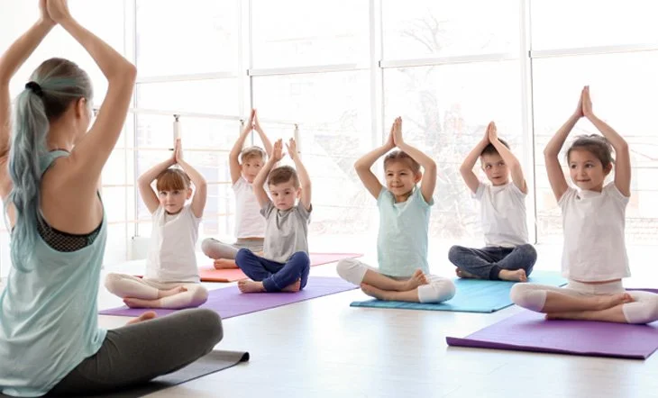 Children participating in a yoga class led by an instructor in a bright room with large windows.