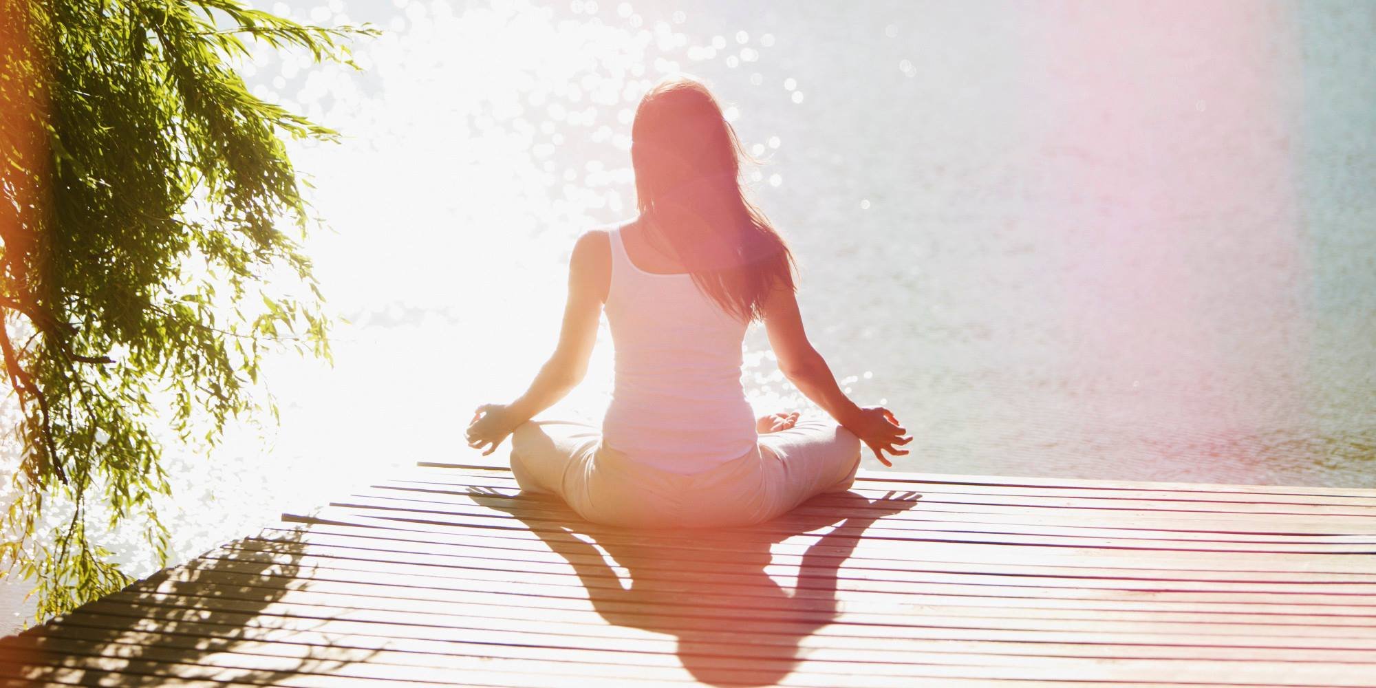 A woman practicing yoga in a seated meditative pose on a wooden dock by the water with sunlight creating a glow.