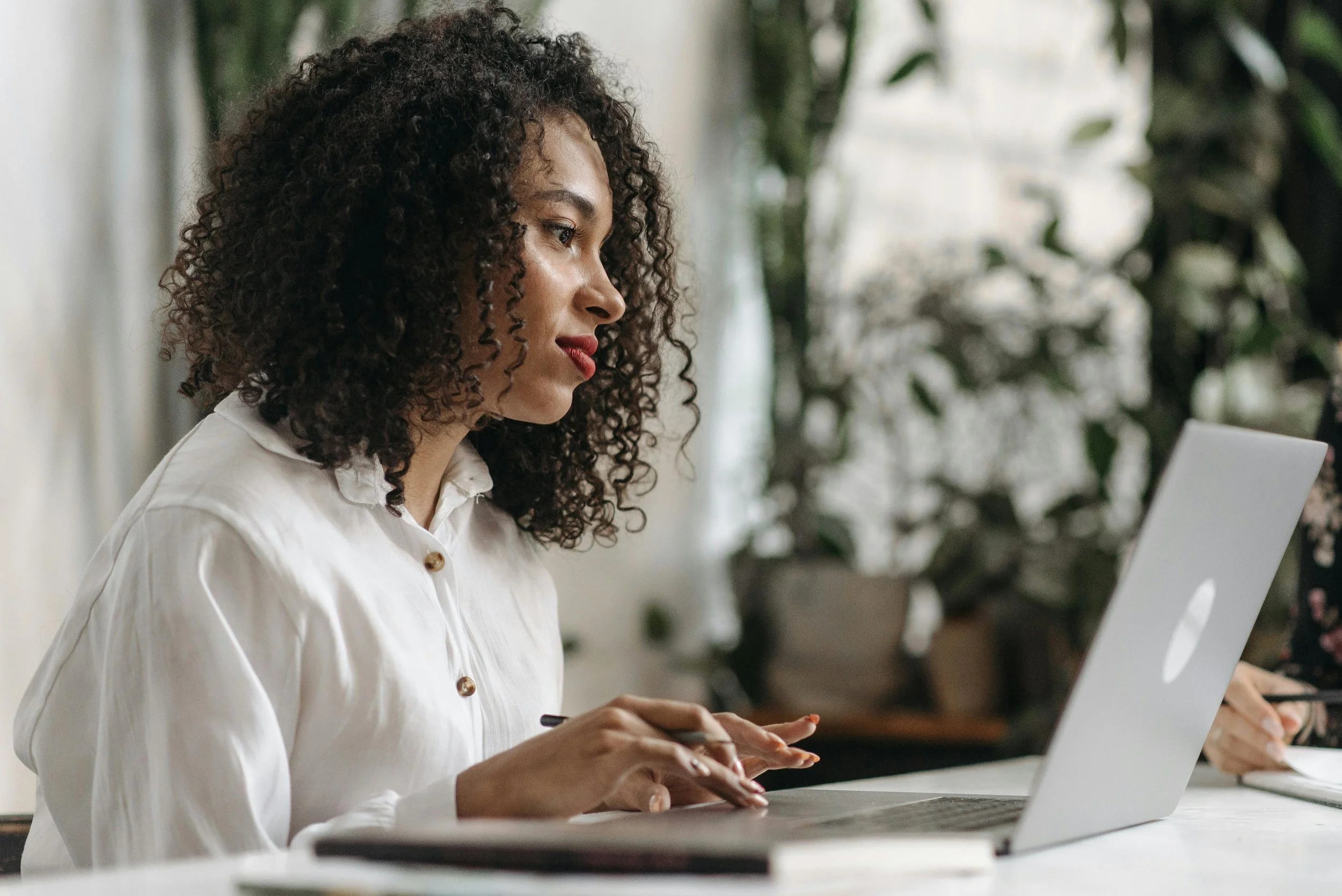 A woman with curly hair wearing a white shirt sitting at a desk working on a laptop.