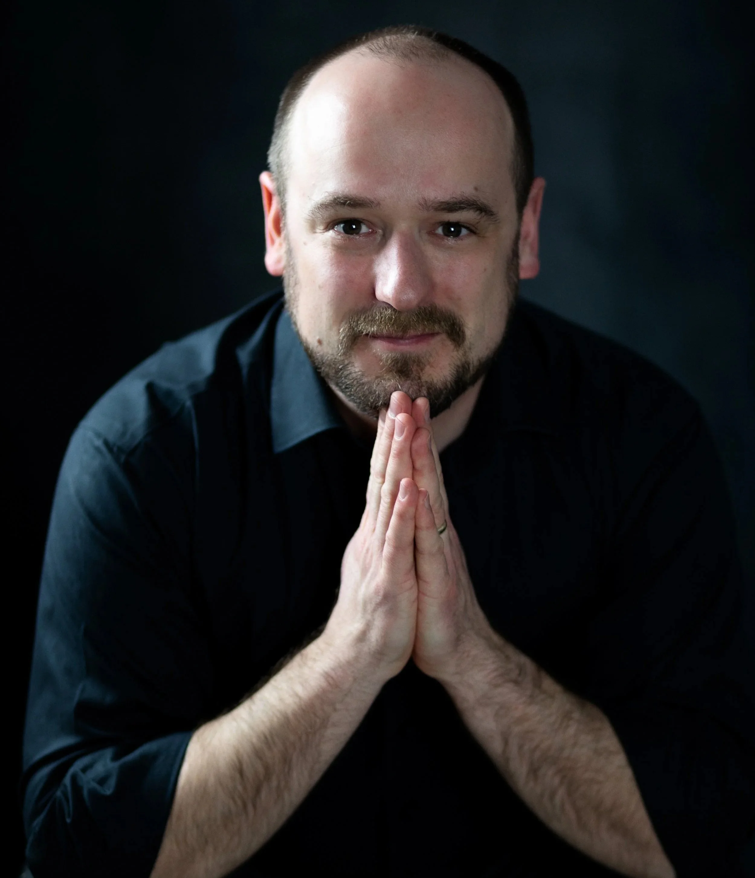A man with short hair and a beard, wearing a black shirt, has his hands pressed together in a prayer-like gesture against a dark background.