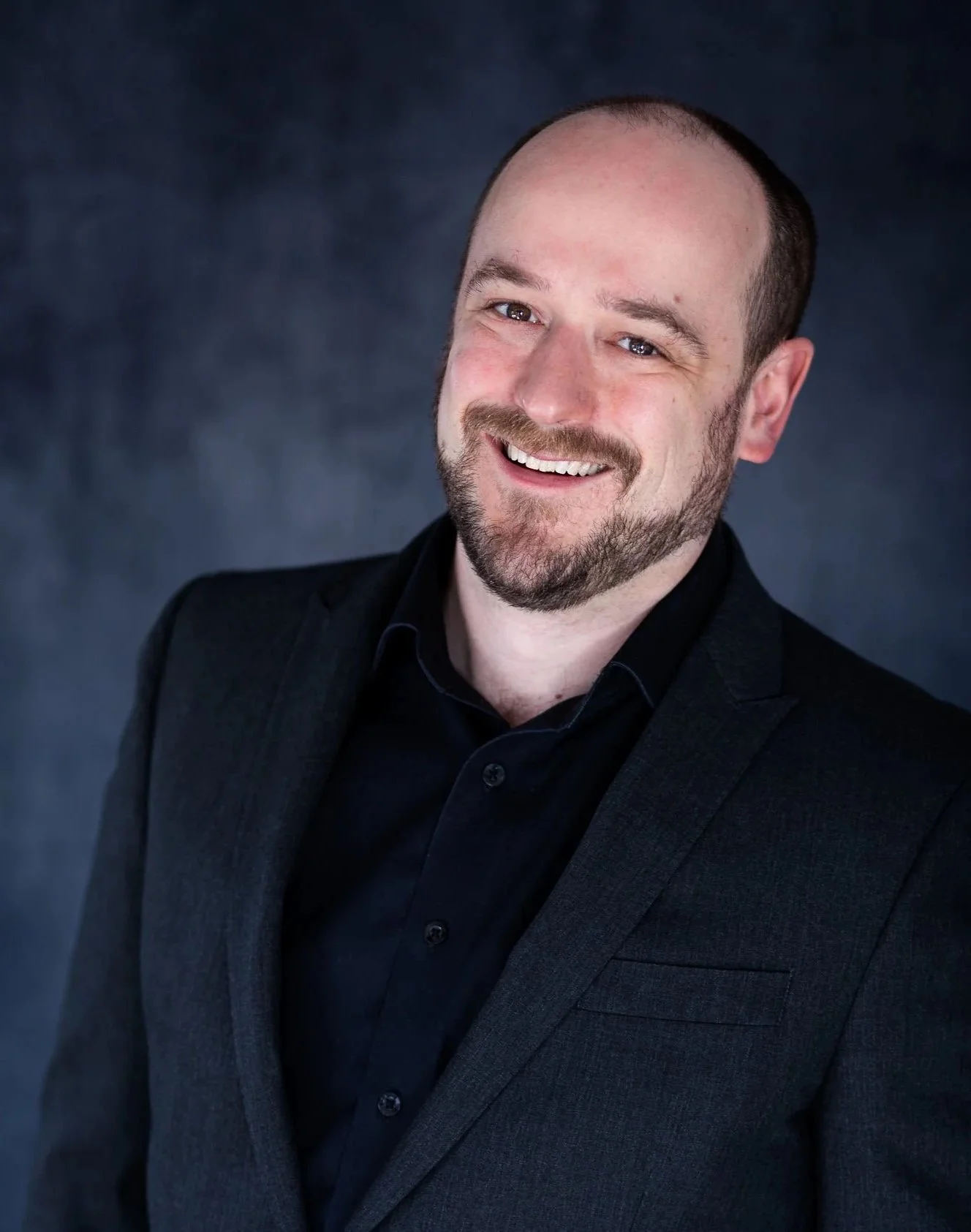 A man with a beard and short hair wearing a black suit and black shirt, smiling, against a dark background.