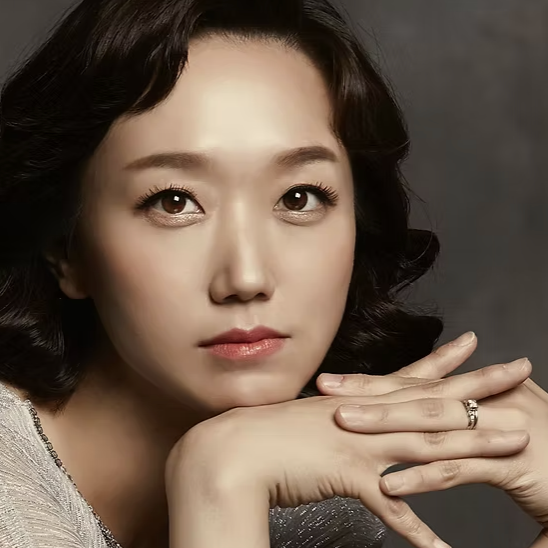 A close-up portrait of an East Asian woman with wavy dark hair, resting her chin on her hands, looking directly at the camera with a neutral expression.