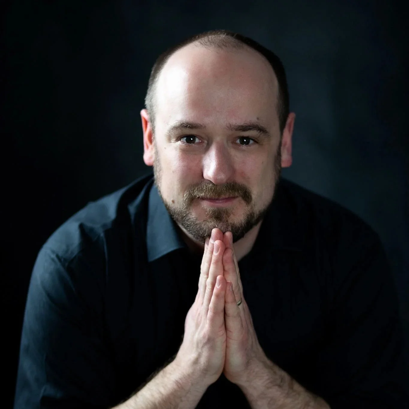Man with a beard, wearing a black shirt, pressing his hands together in a prayer or thinking pose, against a dark background.