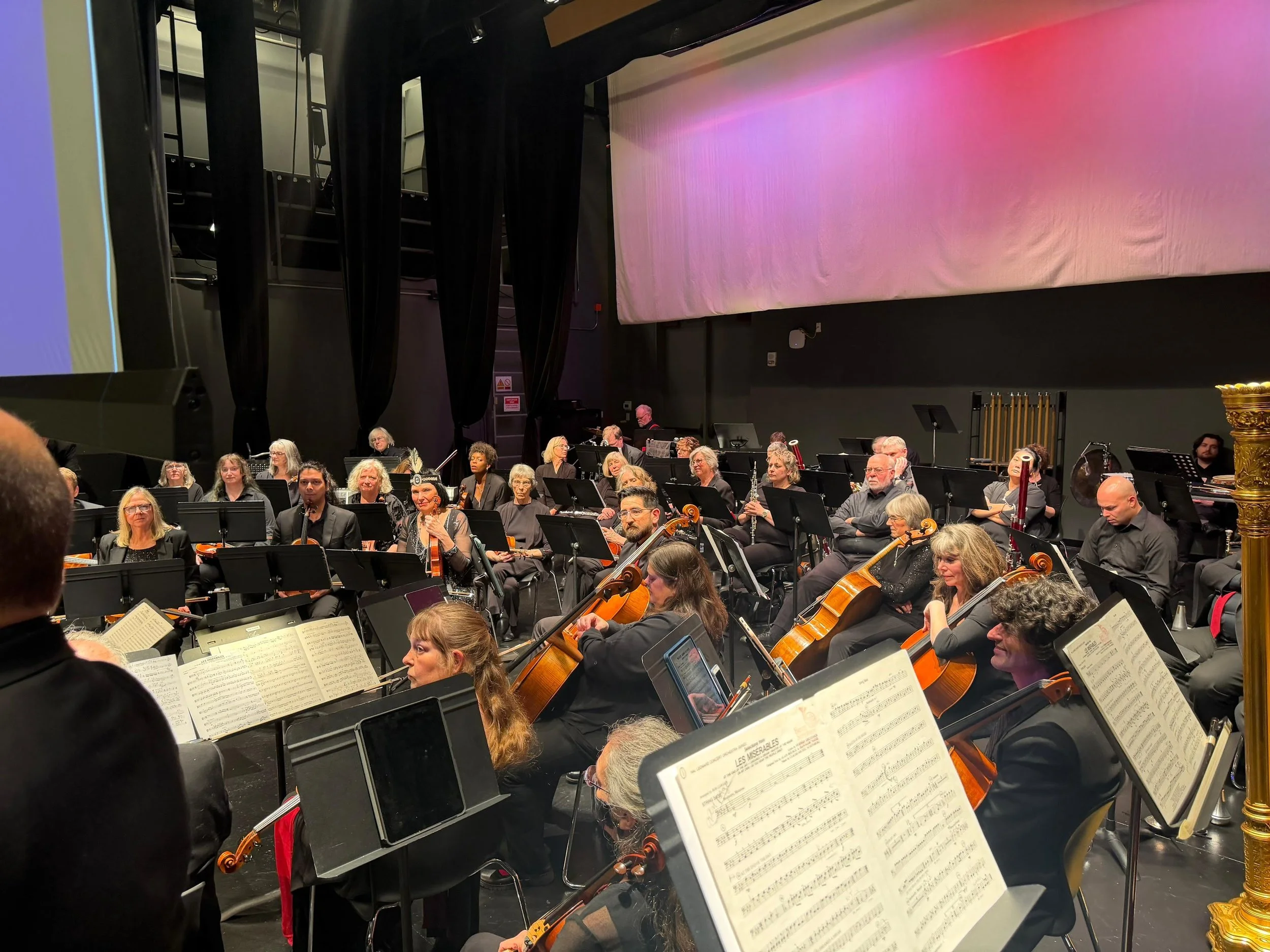 An orchestra performing on stage with musicians playing various instruments, and sheet music on stands in front of them. The stage backdrop displays pink and purple lighting.