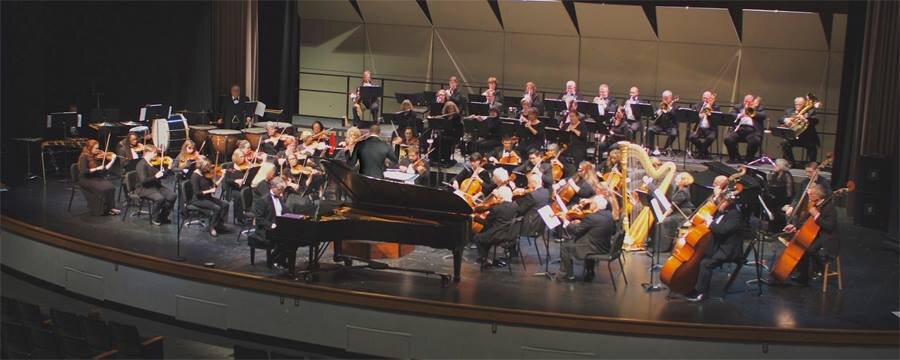 An orchestra performing on stage, with musicians playing various instruments including violins, cellos, clarinets, and a harp, directed by a conductor, in a concert hall.