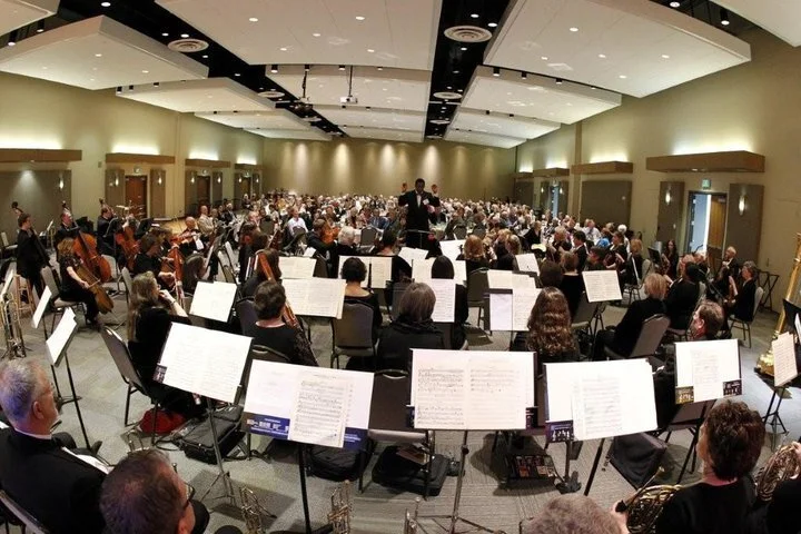 Orchestra performing in a large concert hall with conductor at the center, seated musicians with sheet music, audience in the background.