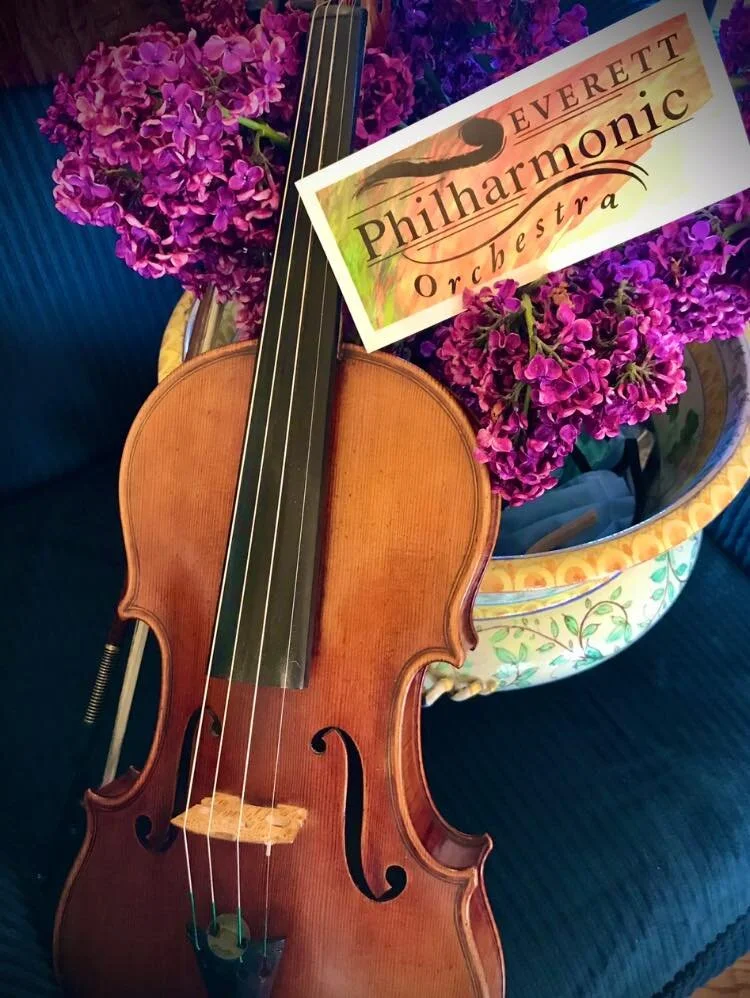 A violin resting on a floral-themed table, surrounded by purple flowers, with a sign that reads 'Everett Philharmonic Orchestra'.