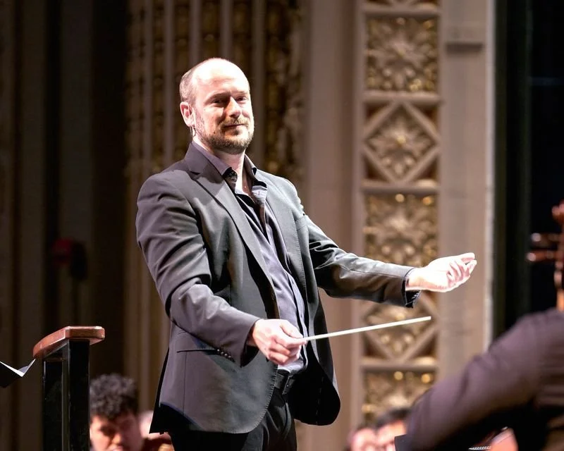 Portrait of a male conductor holding a baton, leading an orchestra during a performance in an ornate concert hall.