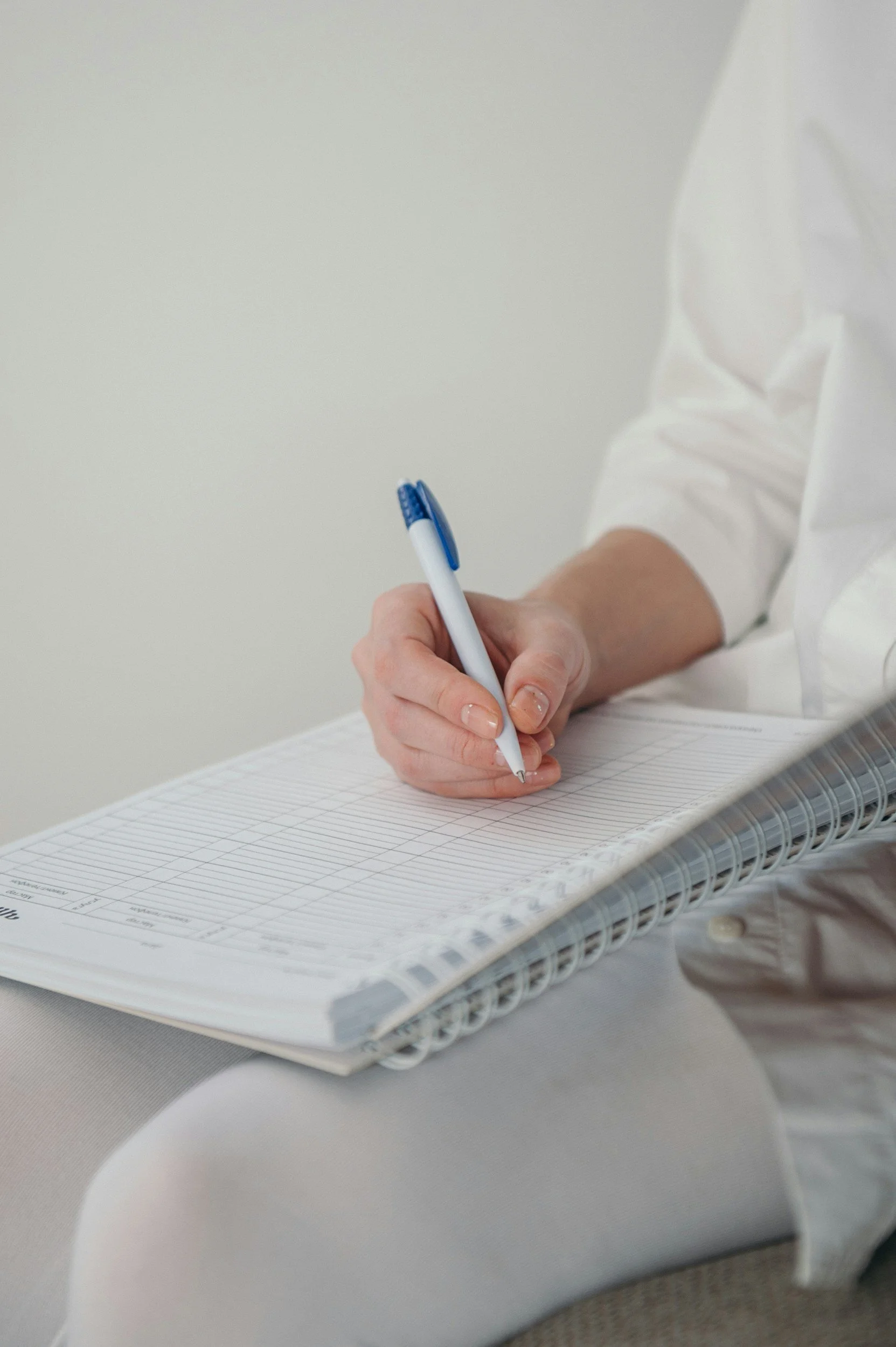 Person in a white shirt writing with a blue pen on a lined planner or calendar on a white surface.