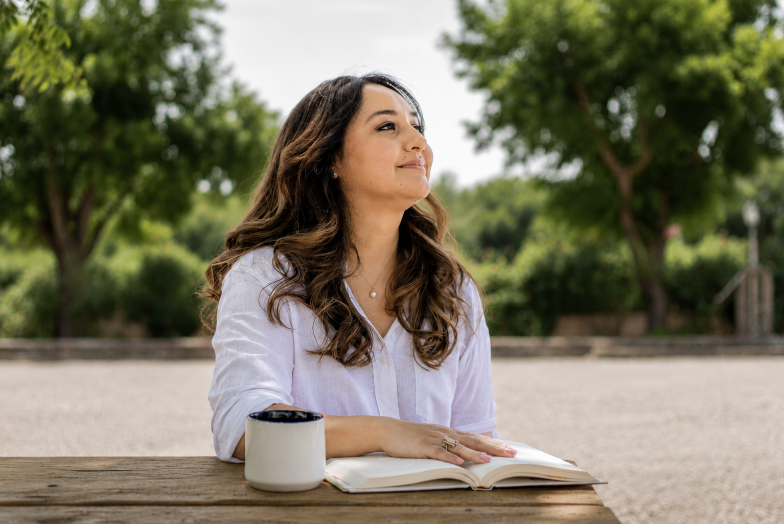 A woman with long wavy brown hair, wearing a white shirt, sits at a wooden outdoor table with a mug and an open book, outdoors with green trees and bright sky.