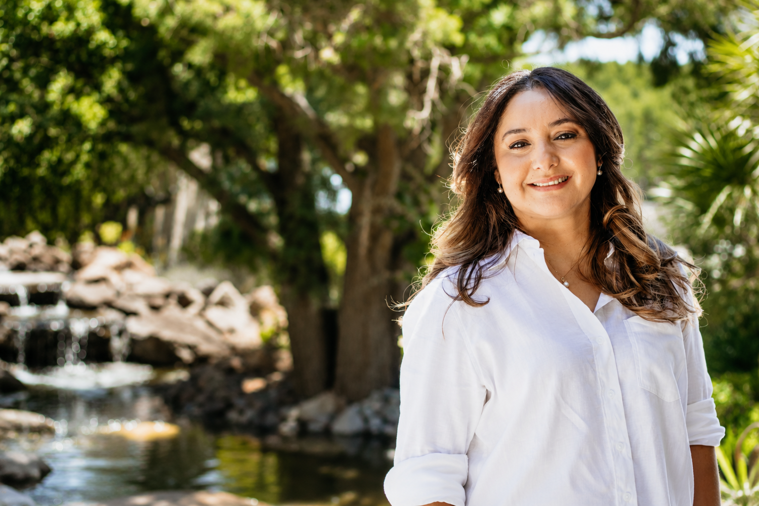 Portrait of a woman smiling outdoors near a small waterfall and trees.