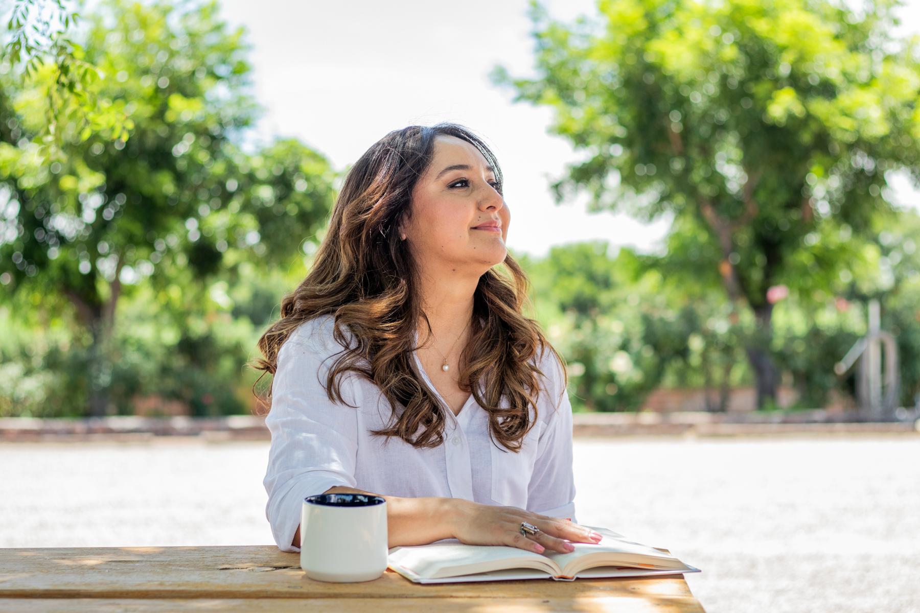 A woman with long brown hair, wearing a white shirt and a pearl necklace, sitting at a wooden table outdoors with trees in the background. She has a white mug and an open notebook in front of her.