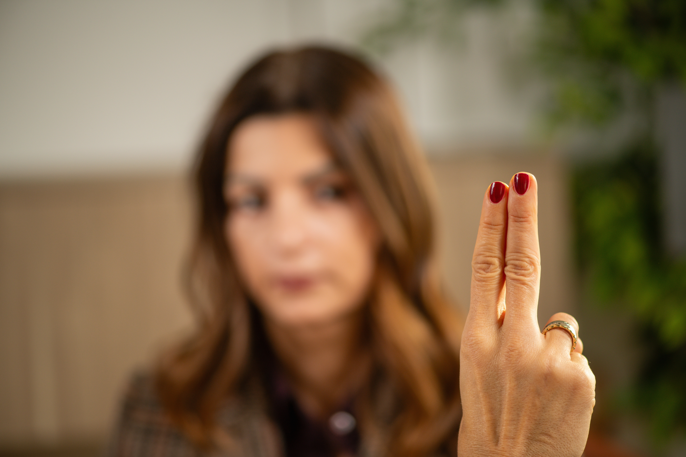 A woman with brown hair, blurred in the background, holding up her index finger with red nail polish and a gold ring, while looking at it.