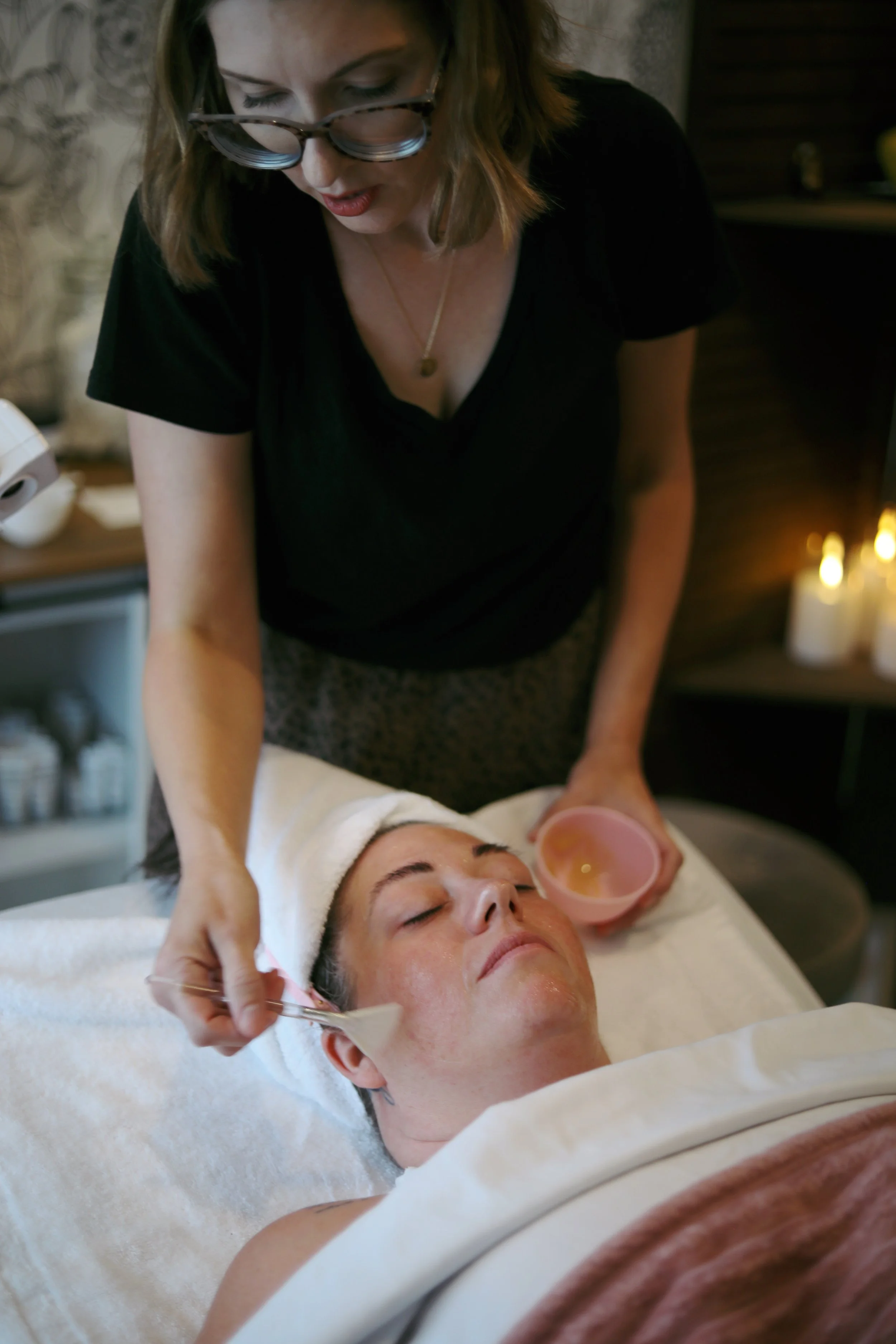 A woman with glasses gives a facial treatment to a woman lying down with his eyes closed in a spa or skincare setting.