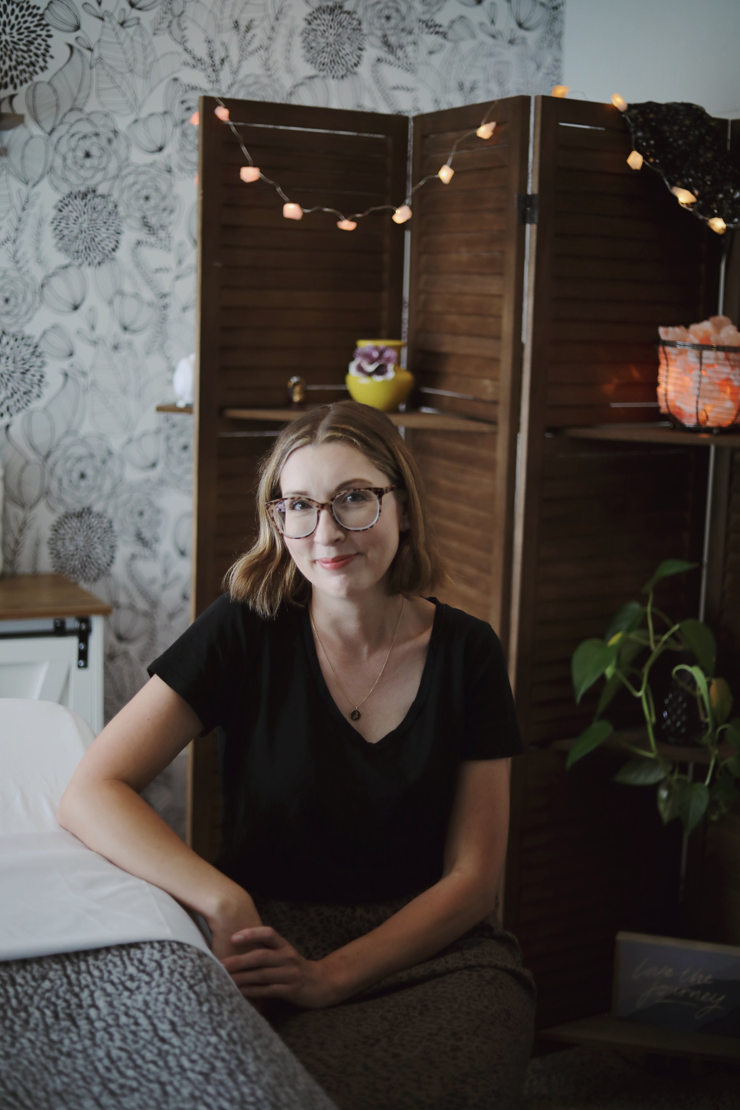 A woman with shoulder-length light brown hair and glasses, wearing a black shirt, sitting on a leopard print cushion in a cozy indoor setting with decorative plants, a folding screen, and string lights.