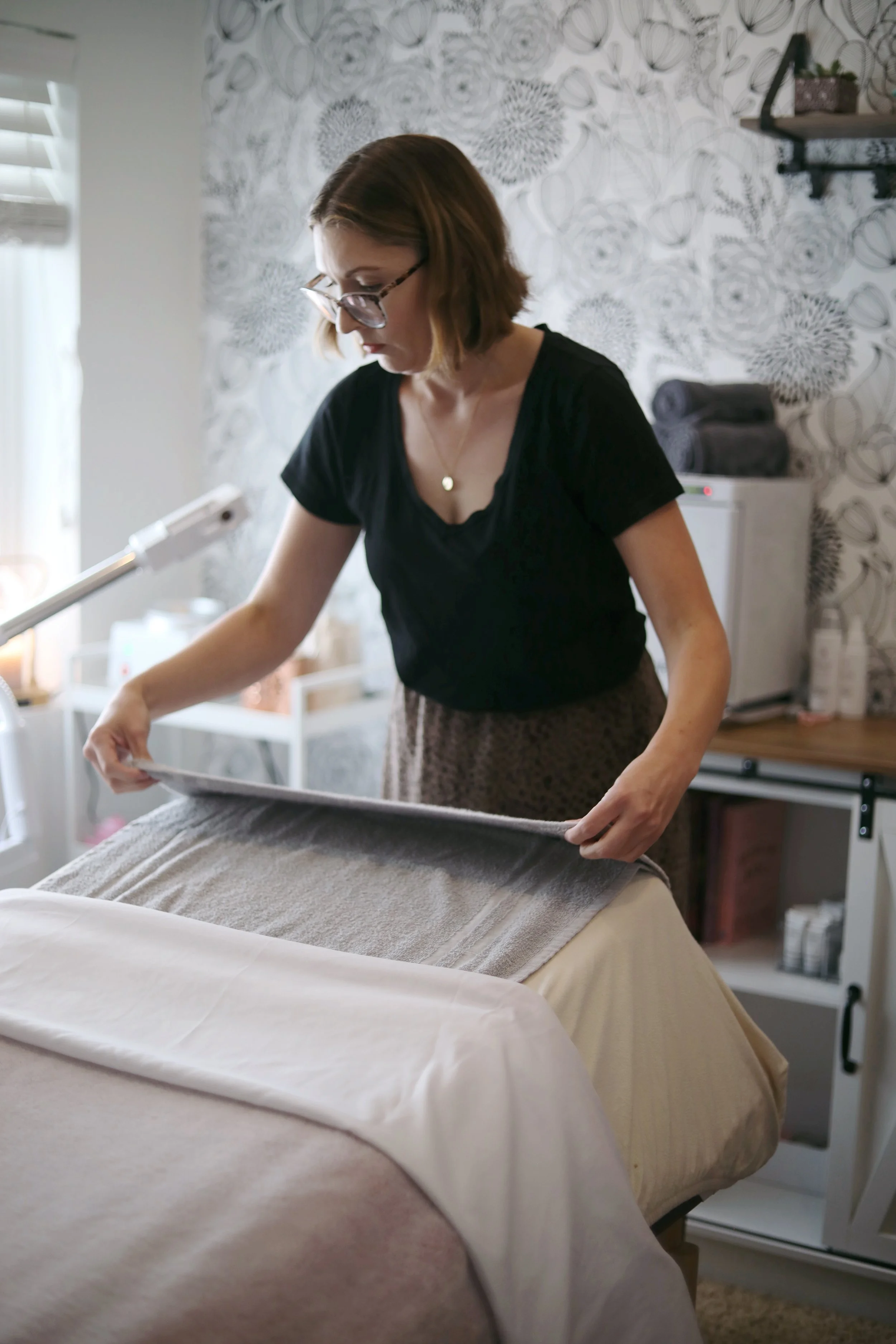 A woman with shoulder-length brown hair, wearing glasses and a black shirt, making a bed with beige sheets in a room with patterned wallpaper and shelf in the background.