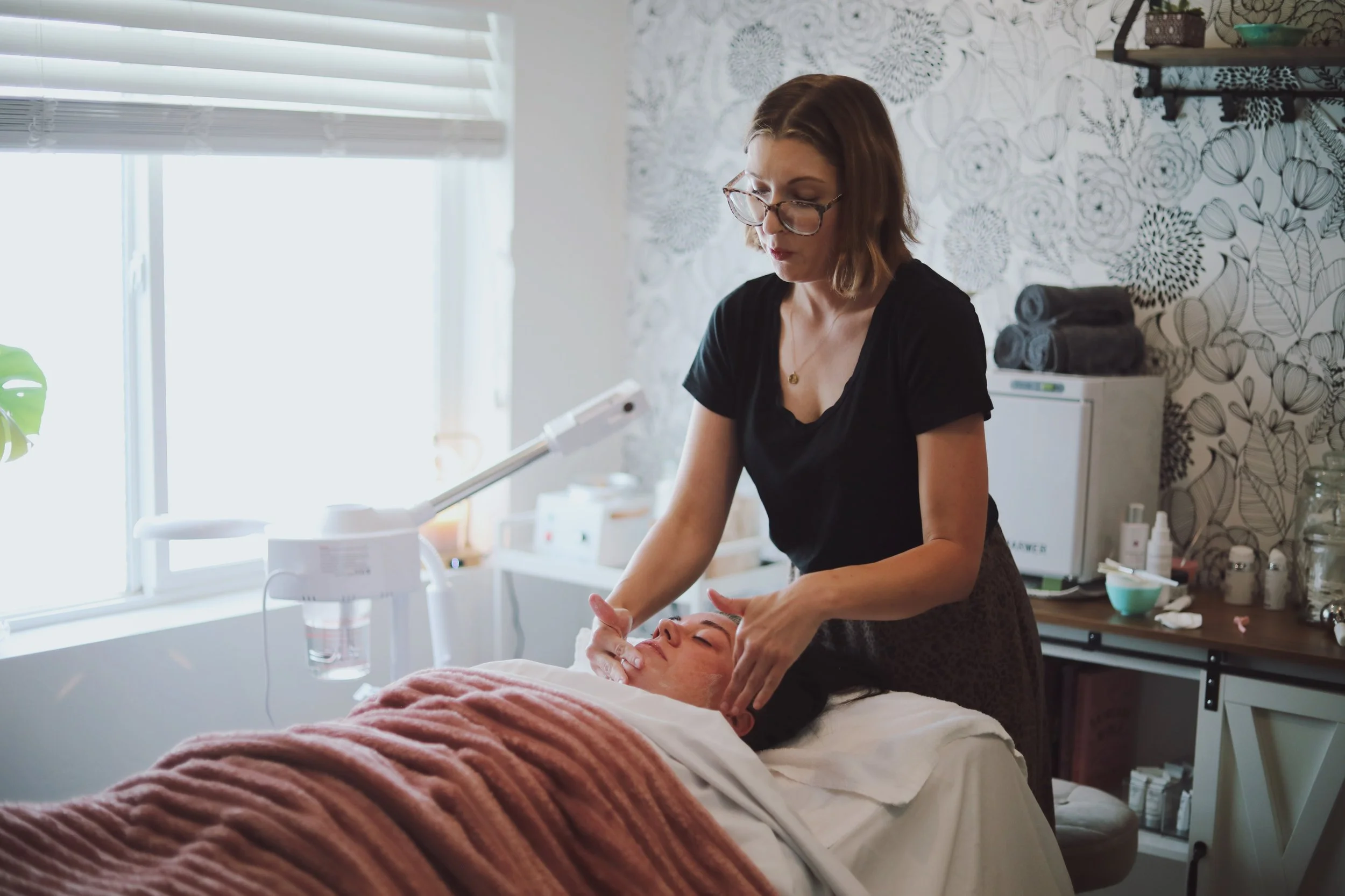 A woman providing a facial treatment to a person lying on a treatment bed in a spa or clinic room with floral wallpaper and shelves with supplies.