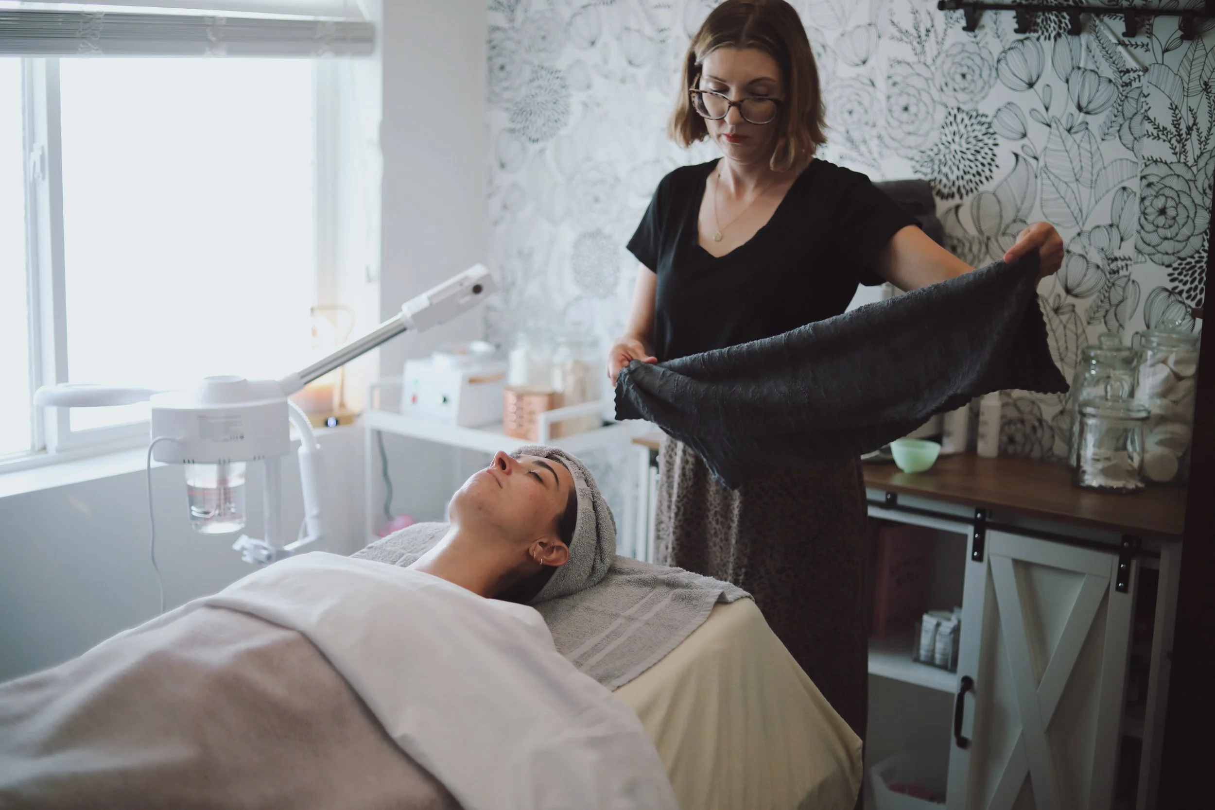 A woman in glasses and black shirt holding a black towel over a woman's face lying in a treatment bed in a room with natural light and patterned wallpaper.