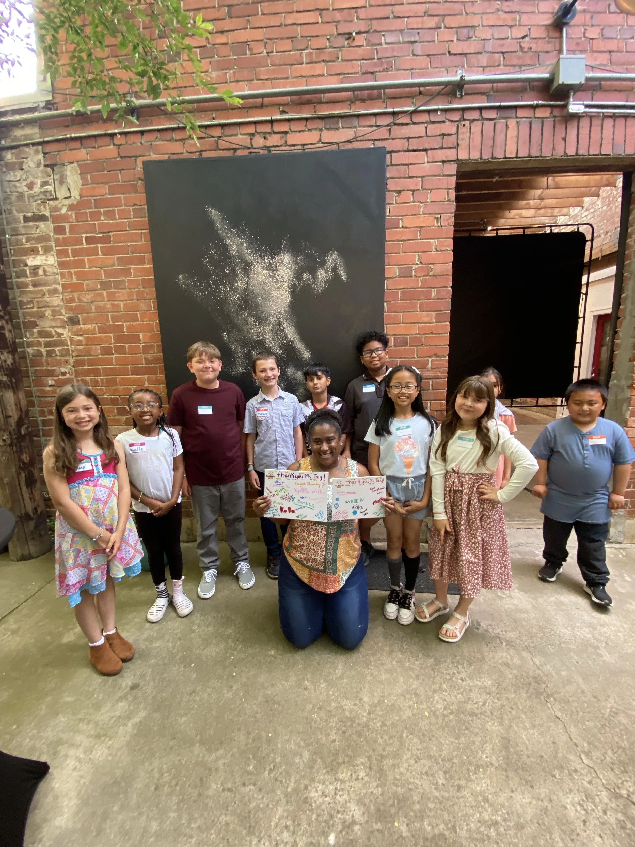 A group of children and an adult woman posing for a photo in an indoor space with brick walls. The woman is kneeling in front, holding up two colorful thank you signs, while the children stand around her, smiling. There is a large black painting or banner with a white abstract splash design on the brick wall behind them.