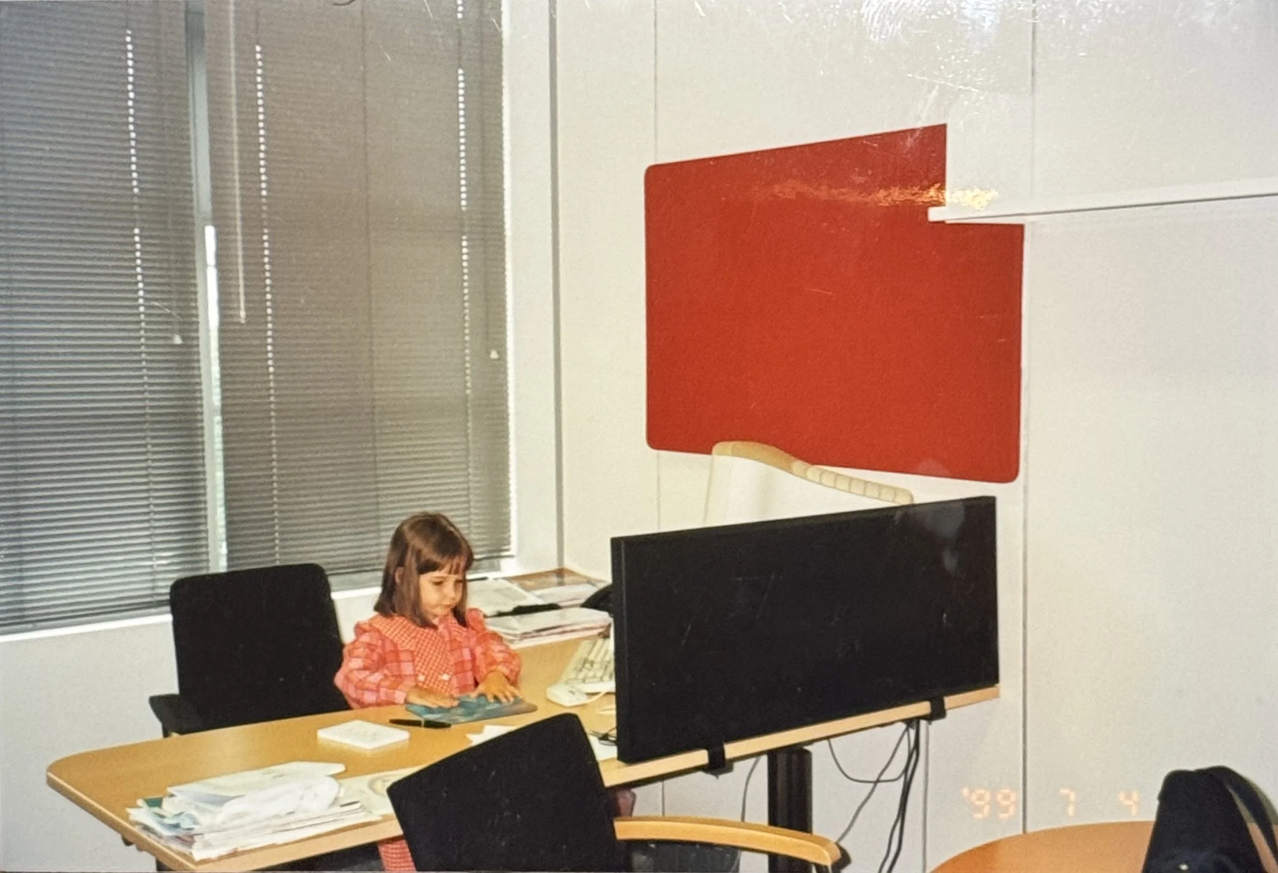 A young girl sitting at a desk with papers in front of her, working on a puzzle, in an office setting with multiple computer monitors and vertical blinds on the window.