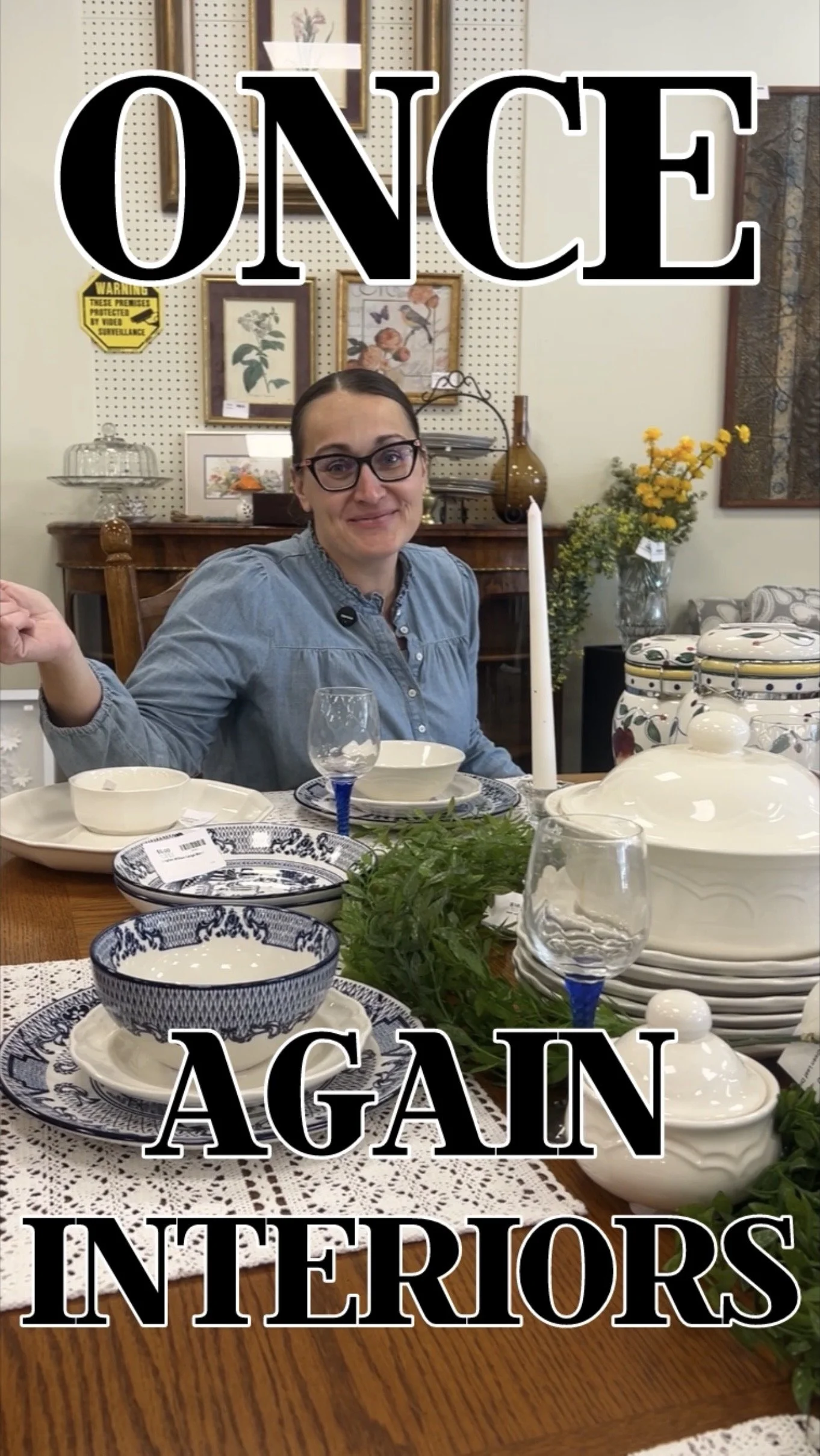 A woman sitting at a dining table with various dishes, plates, glasses, and a candle, inside a home with artwork and decor in the background.