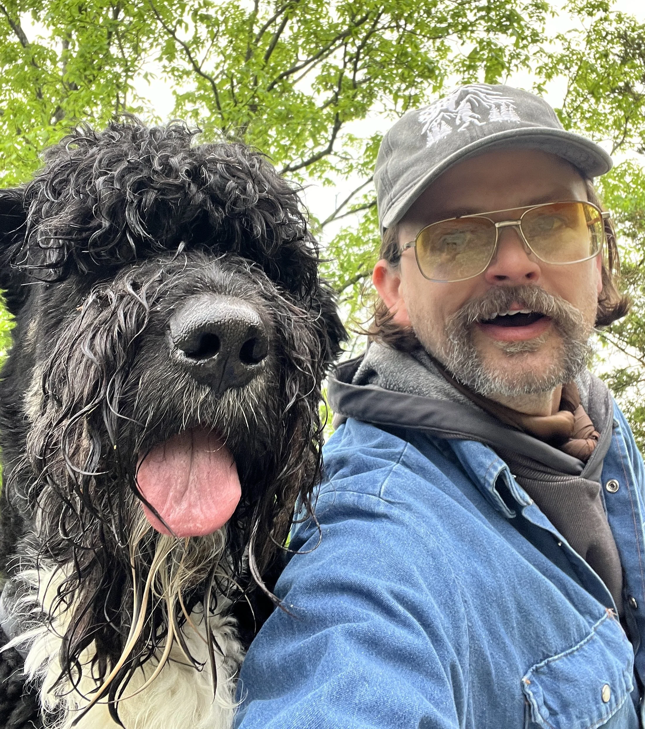 A man with glasses, a gray cap, and a beard taking a selfie outdoors with a large black and white dog with curly wet fur, green leaves and sky in the background.