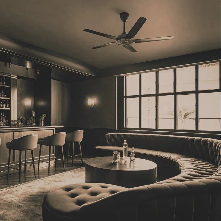 A dimly lit lounge with a curved sectional sofa, a coffee table with bottles and glasses, large windows, a ceiling fan, and a bar area with tall chairs.