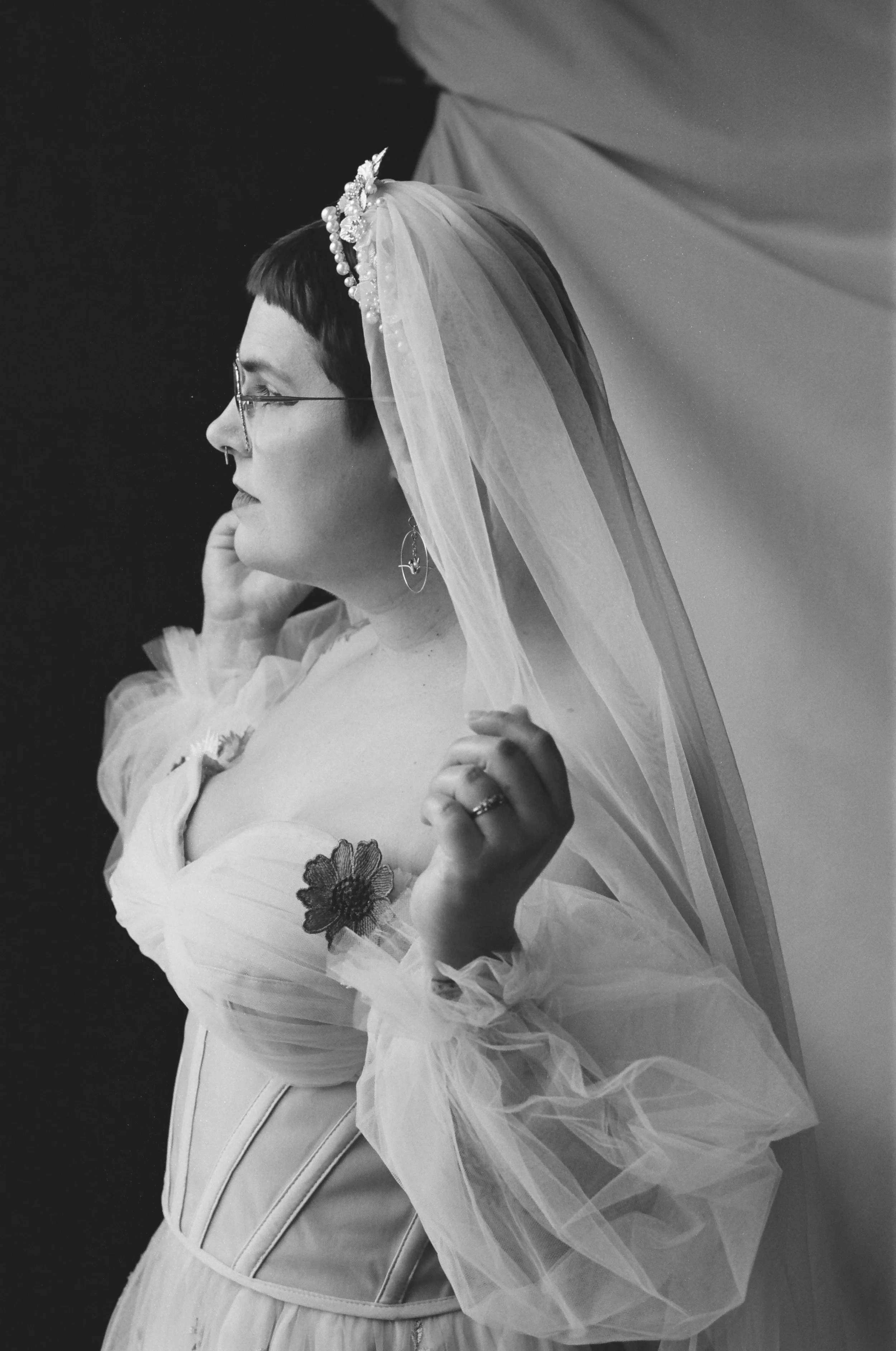 A woman with short dark hair wearing glasses, a veil with pearl and decorative embellishments, a dress with puffy transparent sleeves, floral embroidery on the sleeve, and standing against a dark background.