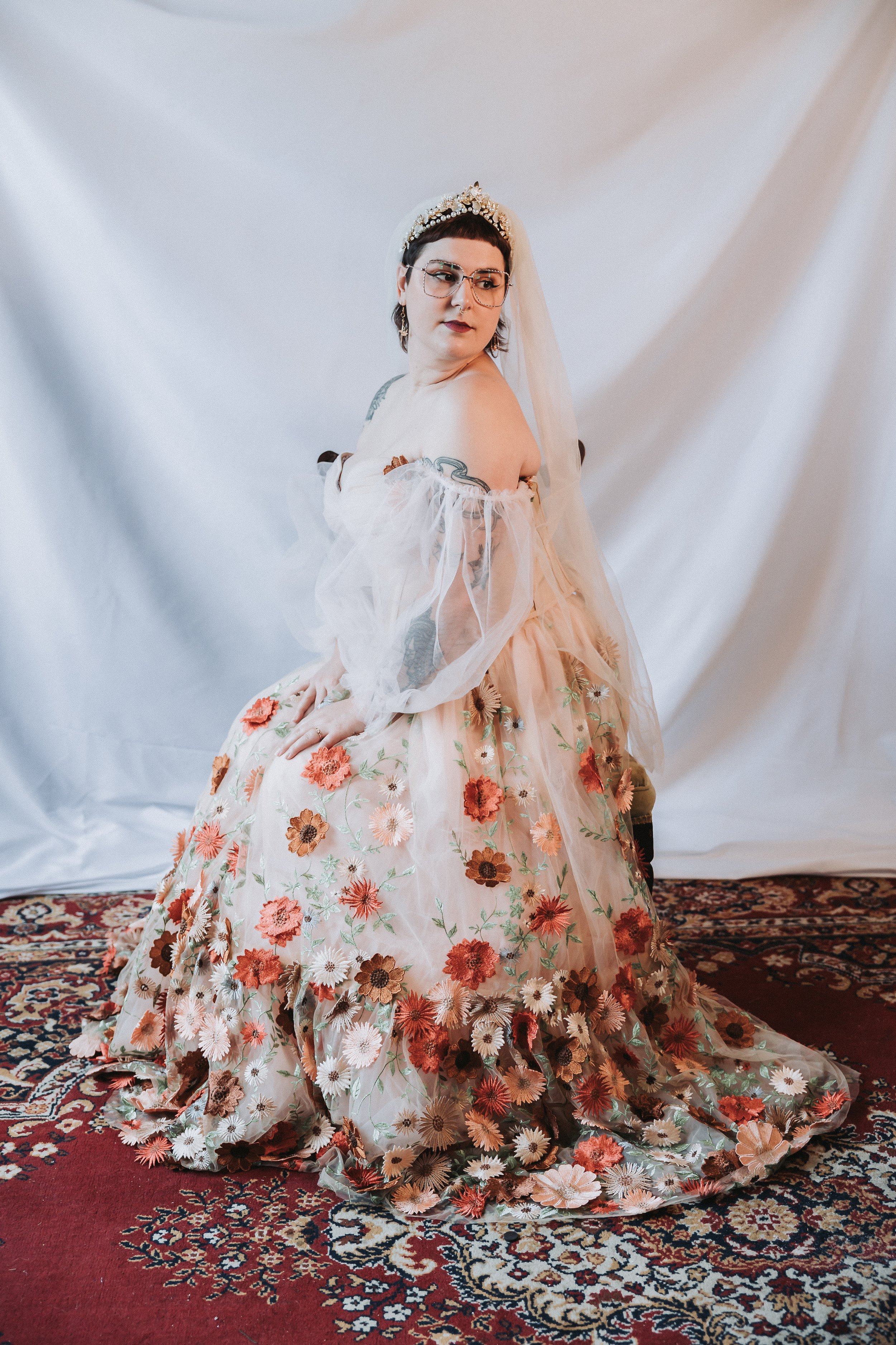 A woman with short dark hair, glasses, and tattoos on her shoulders, wearing an off-shoulder dress with a floral pattern and a veil, standing on a patterned rug with a plain white backdrop.