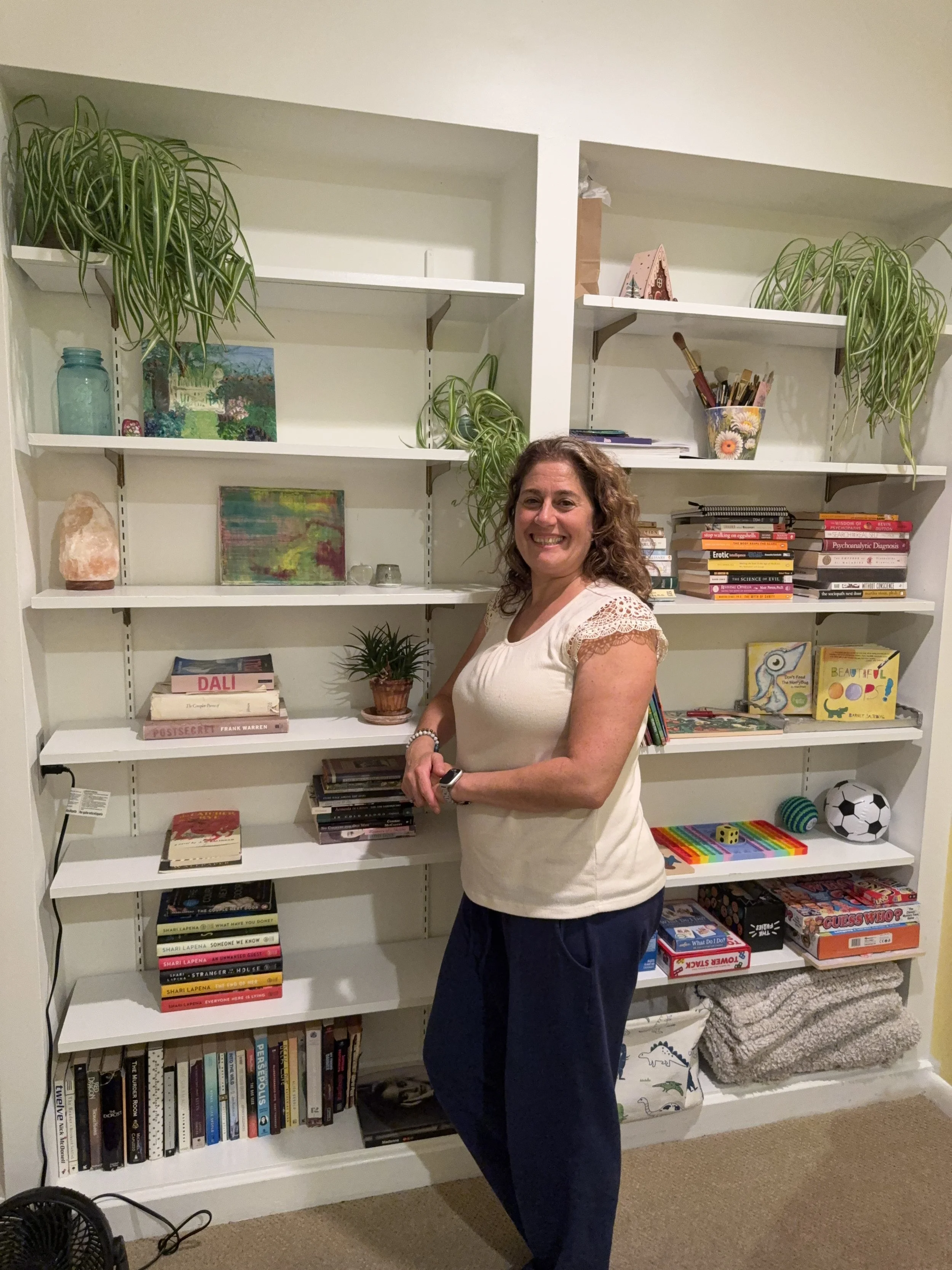 A woman with curly hair smiling at the camera, standing in front of a white bookshelf filled with books, plants, and decorative items in a cozy room.