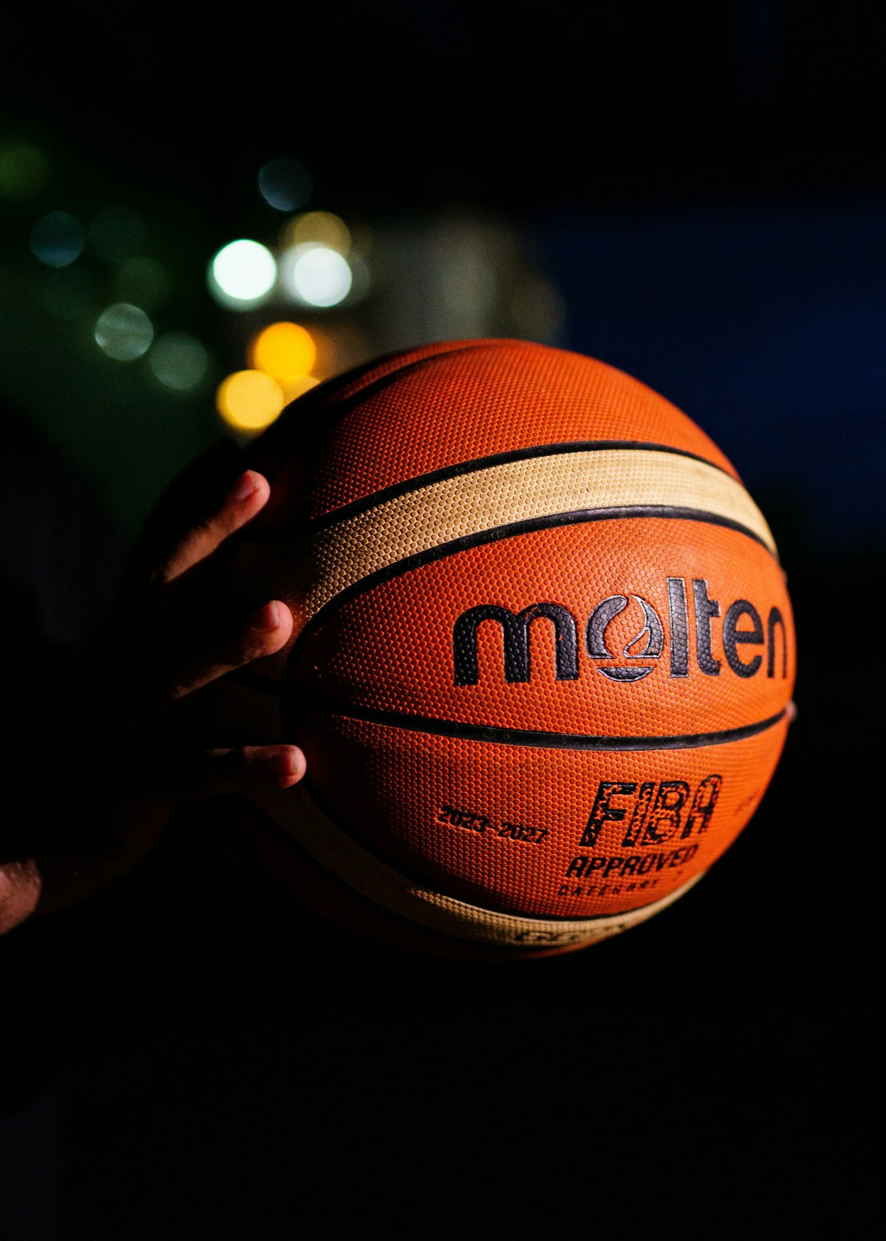 A close-up of an orange Molten basketball being held by a hand in a dark environment with colorful, blurred city lights in the background.