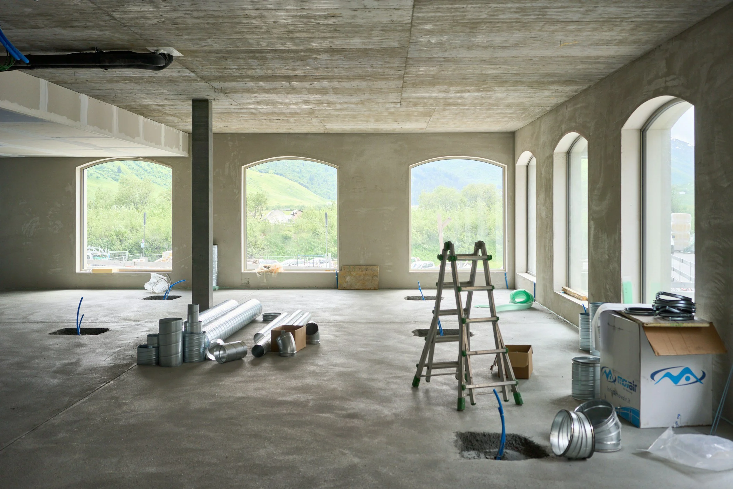 Interior view of an unfinished building under construction, with large windows showing a green landscape outside. Construction materials, pipes, and tools are scattered on the floor, including a ladder, metal ducts, and cans.
